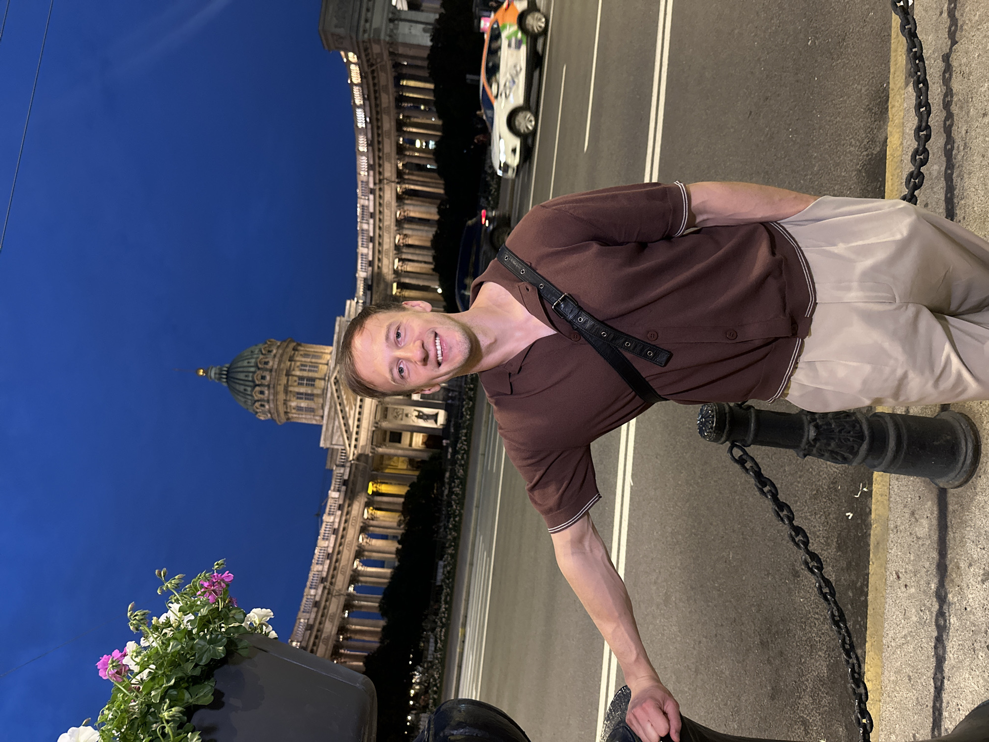 A young Caucasian man with short brown hair and a light complexion is standing in front of the Kazan Cathedral in Saint Petersburg, Russia. He is wearing a short-sleeved brown collared shirt with white trim on the sleeves and light-colored pants. A black strap with silver buckles is slung across his chest. His left arm is resting on a black bollard, and his right hand is in his pocket. He is smiling at the camera.
The Kazan Cathedral, a prominent landmark in Saint Petersburg, is visible in the background. Its large dome and neoclassical colonnade are illuminated, standing out against the deep blue twilight sky. A street with white lane markings is in front of the cathedral, and several cars are driving on it. To the left of the man, a black planter filled with white and pink flowers is mounted on a black pole. A black chain links the bollard to another post on the right. The ground in the foreground is paved with concrete and yellow curb. The overall lighting suggests it is evening or dusk.