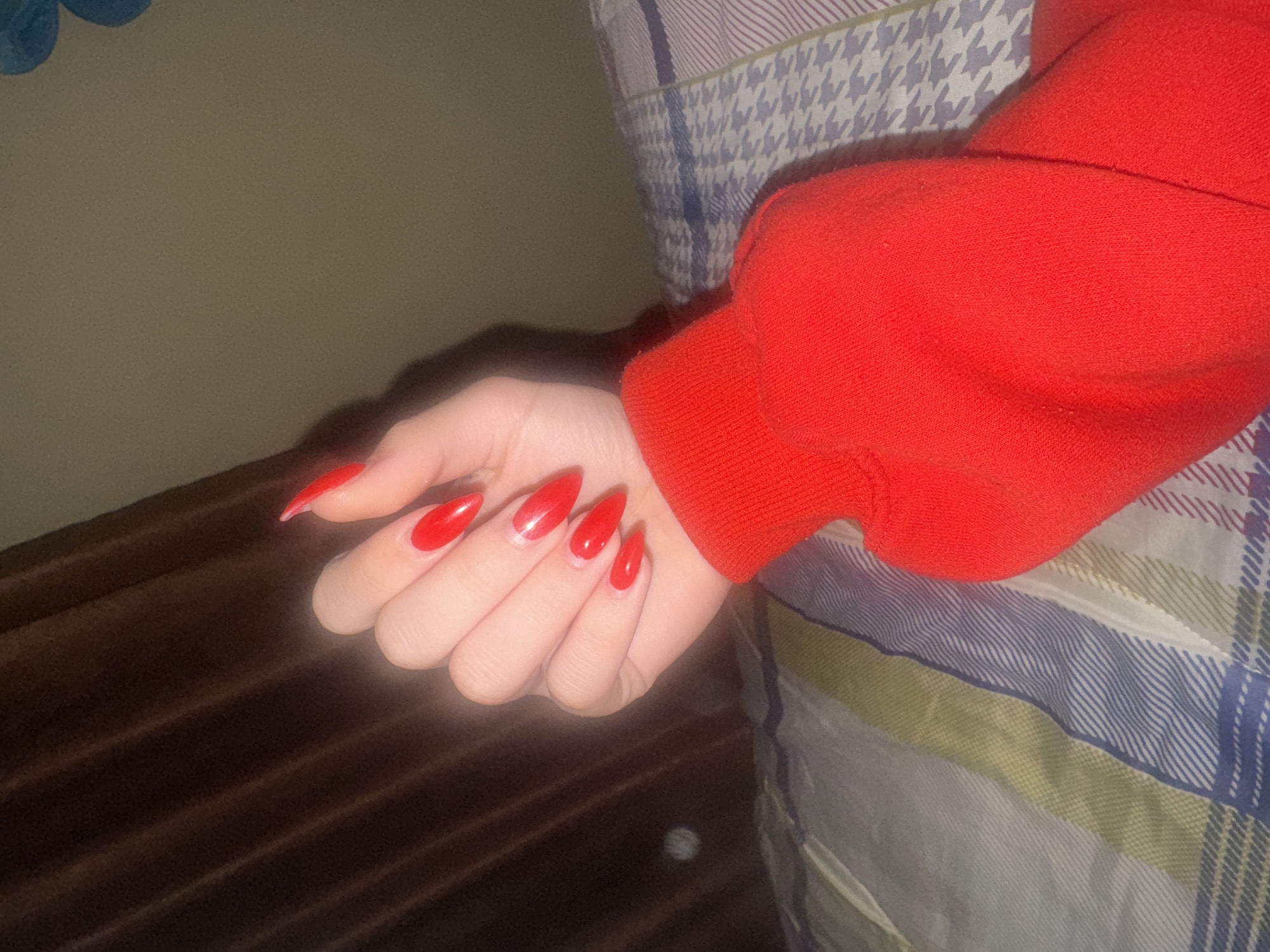 A photograph shows a person's left hand with long, red, almond-shaped fingernails. The person is wearing a red sweater or sweatshirt. The hand is resting on a surface; partially visible is a plaid or striped pattern that may be bedding or a blanket. The background is partially visible with the appearance of a brown textured surface to the left and an off-white or light-colored wall in the background. The location of Lehchevo, Bulgaria is not discernible from the image.