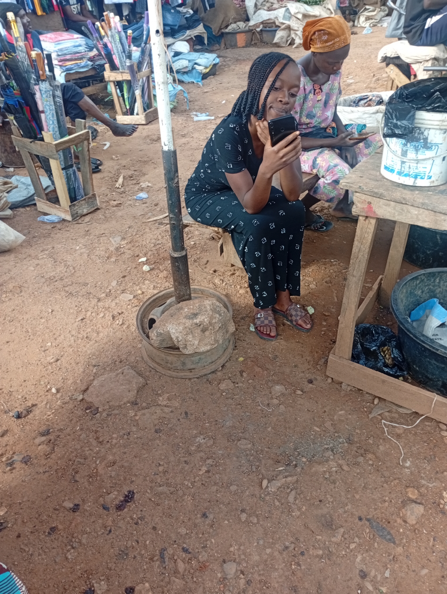 This media file captures a scene in Rafin Par, Nigeria, likely during the daytime given the lighting. Two young women are the primary subjects, seated on a wooden bench or structure. The woman in the foreground, on the left, is wearing a black dress with a white patterned print and brown checkered sandals. She is actively engaged with her smartphone, holding it in her hands and looking down at the screen with a focused expression. Her braided hair is pulled back. To her right sits another young woman wearing a light-colored, patterned dress and an orange headscarf. She is also looking down, seemingly engaged with her own phone, though her hand is partially obscured.
The setting appears to be an outdoor market. The ground is made of reddish-brown dirt with scattered stones and debris. Behind the women, various market stalls are visible, with stacks of folded fabrics and what appear to be umbrellas in wooden crates. A metal pole, possibly part of a stall structure, stands vertically in the foreground, secured by a large stone placed inside a metal rim. To the right of the women, a white bucket with blue and white lettering is visible, along with a black plastic tub containing some items wrapped in blue and white plastic. The overall atmosphere suggests a busy, perhaps a bit dusty, market environment. The focus on the smartphones indicates a common modern activity taking place even in what appears to be a traditional market setting.