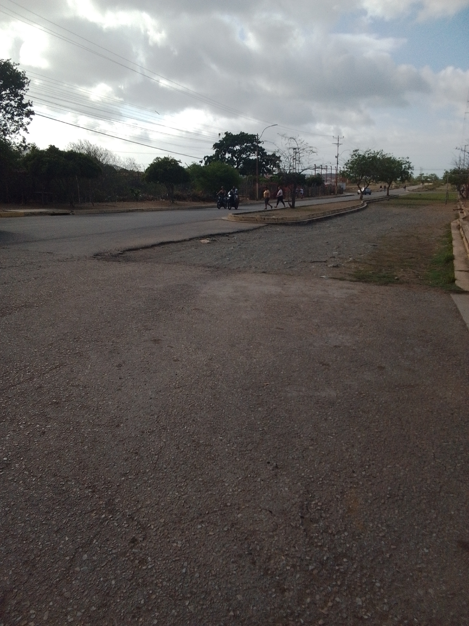 A street scene in Altagracia, Venezuela, under a cloudy sky. The foreground shows a dark asphalt road with a gravelly texture. In the mid-ground, a paved road curves to the left, bordered by a concrete curb and a strip of dry grass. On the left side of the paved road, several people are walking, and a motorcycle is visible. Further back, trees and power lines are present, along with more vegetation and distant structures. The right side of the image features a sidewalk with a concrete curb and a grassy verge.