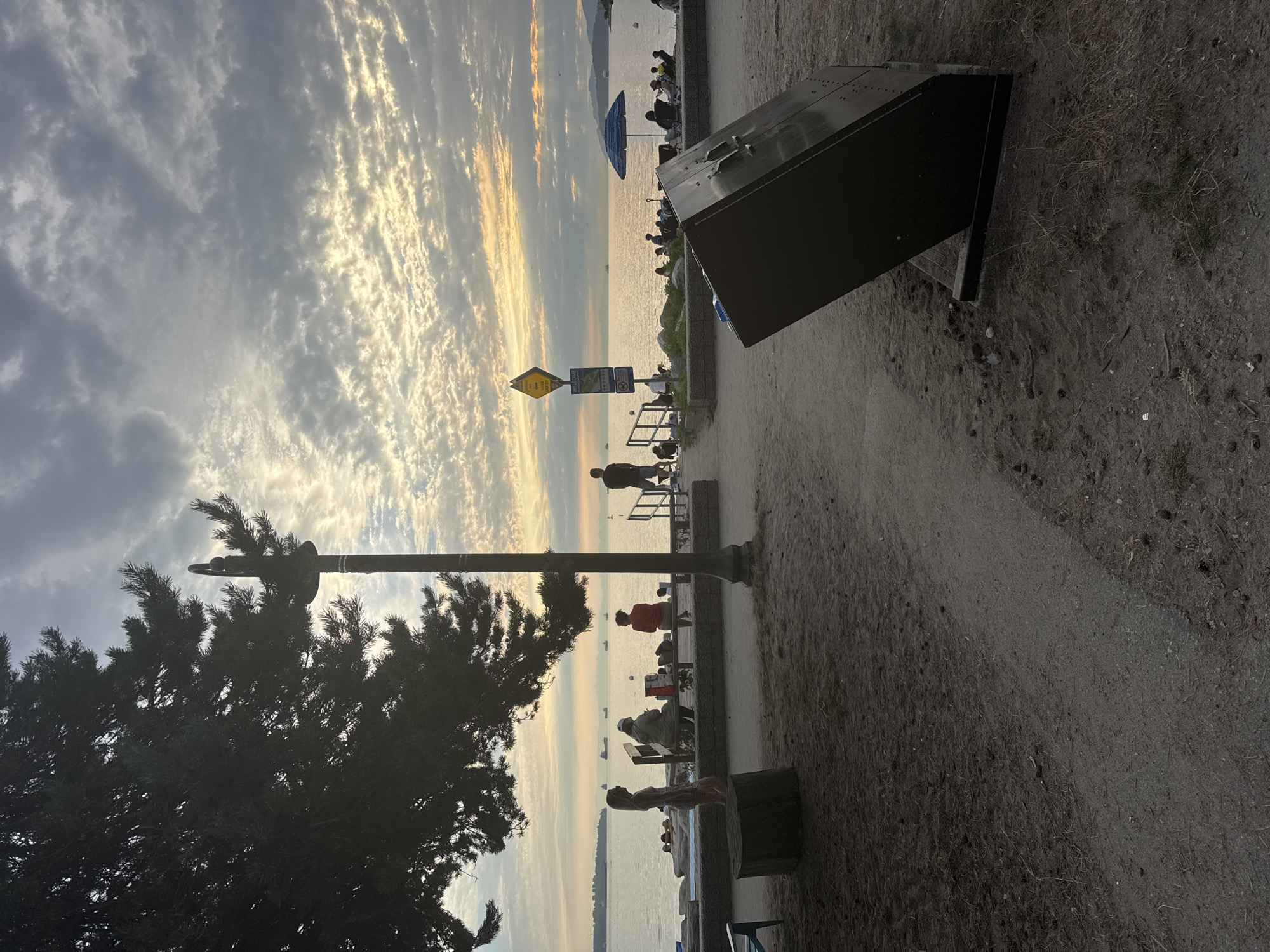 At sunset, people are relaxing on a sandy beach in West Vancouver, Canada. In the foreground, a large black metal object is tilted on the sand. A paved path leads to a low stone wall separating the sandy area from the beach, where a few people are seated on benches or lying on towels. Further along the beach, a blue umbrella provides shade for several people in lawn chairs. The ocean stretches to the horizon, dotted with several ships, and mountains are visible in the distance. The sky is filled with dramatic, streaky clouds illuminated by the setting sun. A tall lamp post stands near the center of the frame, and a large tree casts a shadow on the left side of the image. A woman in a light-colored dress stands near a tree stump on the left.