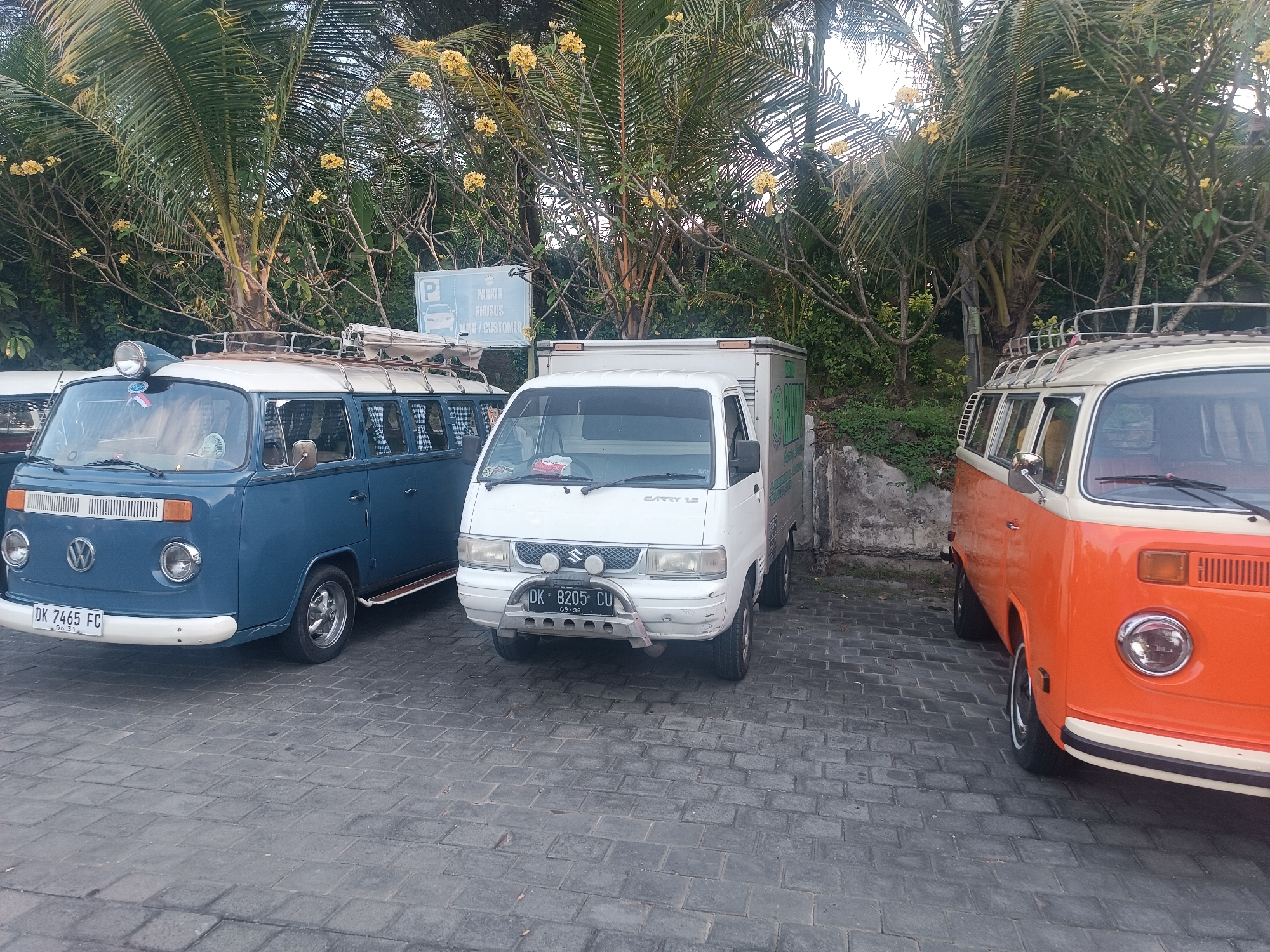The image captures a frontal view of three vehicles parked on a paved surface. The leftmost vehicle is a blue Volkswagen van with a white roof, featuring a roof rack. To its right is a white Suzuki Carry pickup truck with a white cargo box. The rightmost vehicle is an orange and white Volkswagen van, also with a roof rack, showing its front and side profile. Palm trees and other vegetation are visible in the background, with a sign that includes a parking symbol and text in Indonesian. The scene appears to be an outdoor parking area, possibly in a tropical or subtropical location, given the presence of palm trees and flowering plants. The license plates of the vehicles indicate they are registered in Indonesia. The blue van has a license plate reading "DK 7465 FC", the white truck reads "DK 8205 CU", and the orange van's license plate is partially obscured. The setting is consistent with a location in the city of Kediri, Indonesia, or a similar urban environment with lush tropical vegetation.