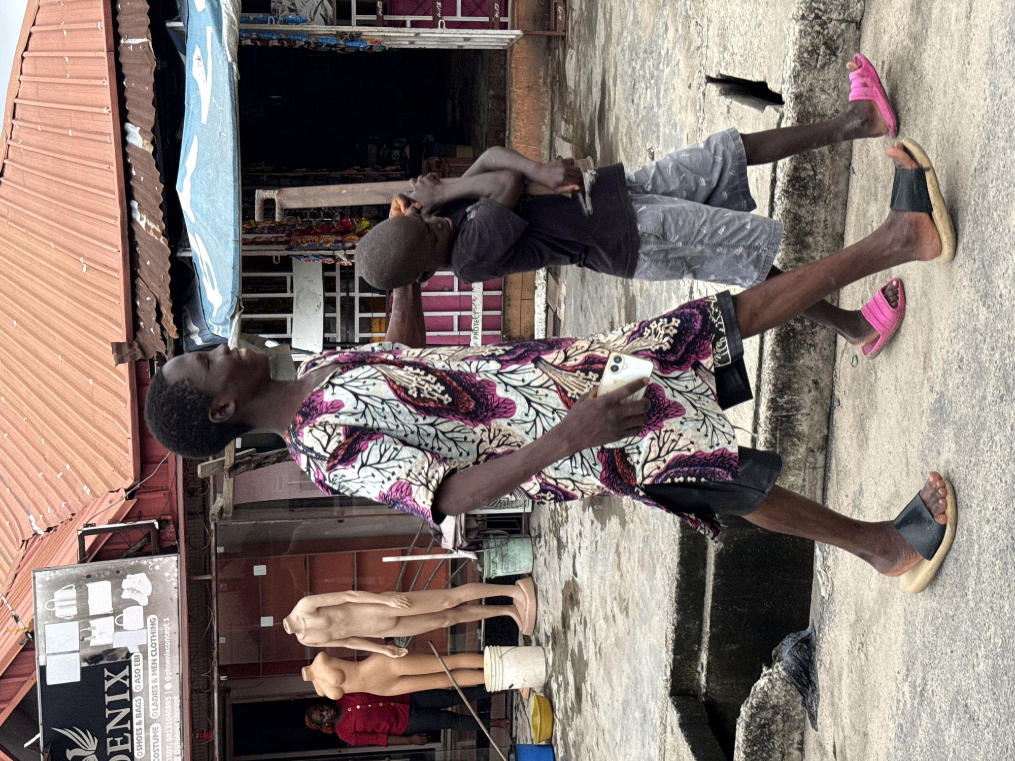 The scene is set in Ado, Nigeria, alongside a street. An adult male stands holding a smartphone, wearing a white patterned shirt with purple and black, and black shorts. His feet are clad in a black sandal on one foot and a pink sandal on the other. Beside him is a child in a black shirt and patterned gray shorts, also wearing pink sandals. Behind them is a commercial building with a sign that reads "DENIX," advertising shoes, bags, costumes, and clothing. Two mannequins stand in front of the building, and a woman in a red shirt can be seen inside. Various containers, including buckets and a barrel, are present near the mannequins. The street and sidewalk show signs of wear.