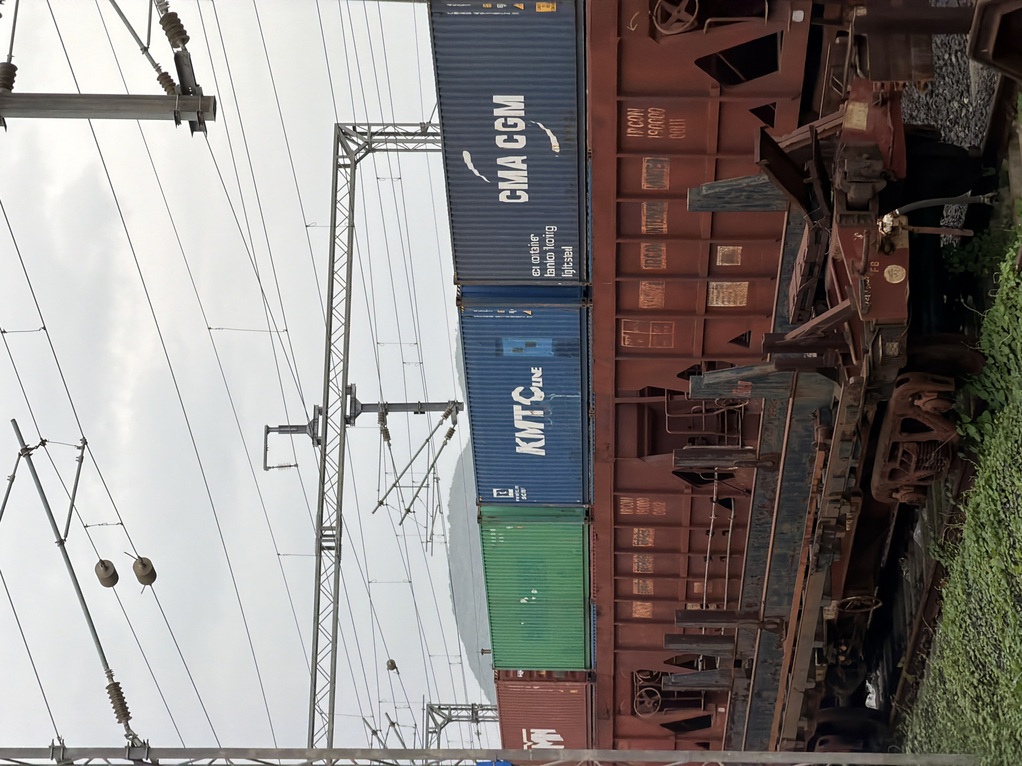 The image depicts a freight train carrying containers, parked on railway tracks in Saphale, India. The train consists of several open-top railcars loaded with shipping containers. In the foreground, a rusty, multi-level railcar is visible, with various metal components and what appears to be a coupling system. Behind this, three stacked shipping containers are prominently featured: a red container on the left, a green container in the middle, and a blue container on the right. The blue container bears the white logo and text "CMA CGM". Above the train, overhead power lines and supporting infrastructure for an electrified railway system are clearly visible against an overcast sky. The ground beneath the tracks is covered with lush green vegetation. No people are present in the frame. The scene appears to be an outdoor industrial or transportation hub, likely a railway yard or loading facility within Saphale.