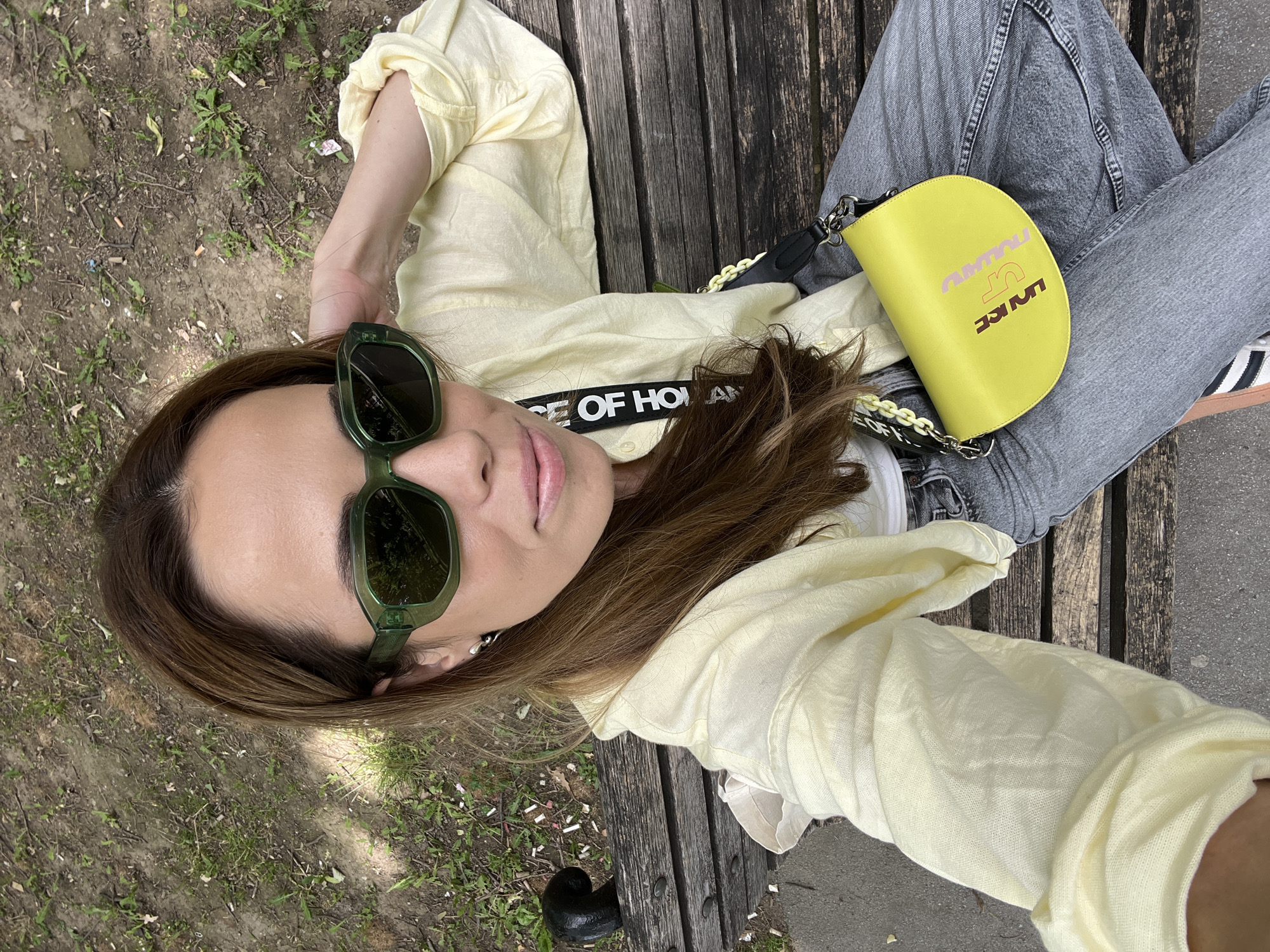 The image is an overhead shot of a woman lying on a wooden park bench. She is wearing green sunglasses, a light yellow, unbuttoned, long-sleeved shirt, grey jeans, and is carrying a yellow handbag. Part of a black and white shoe is also visible. The bench is located in a grassy area, and a concrete path borders it. The image was taken in Belgrade, Serbia.