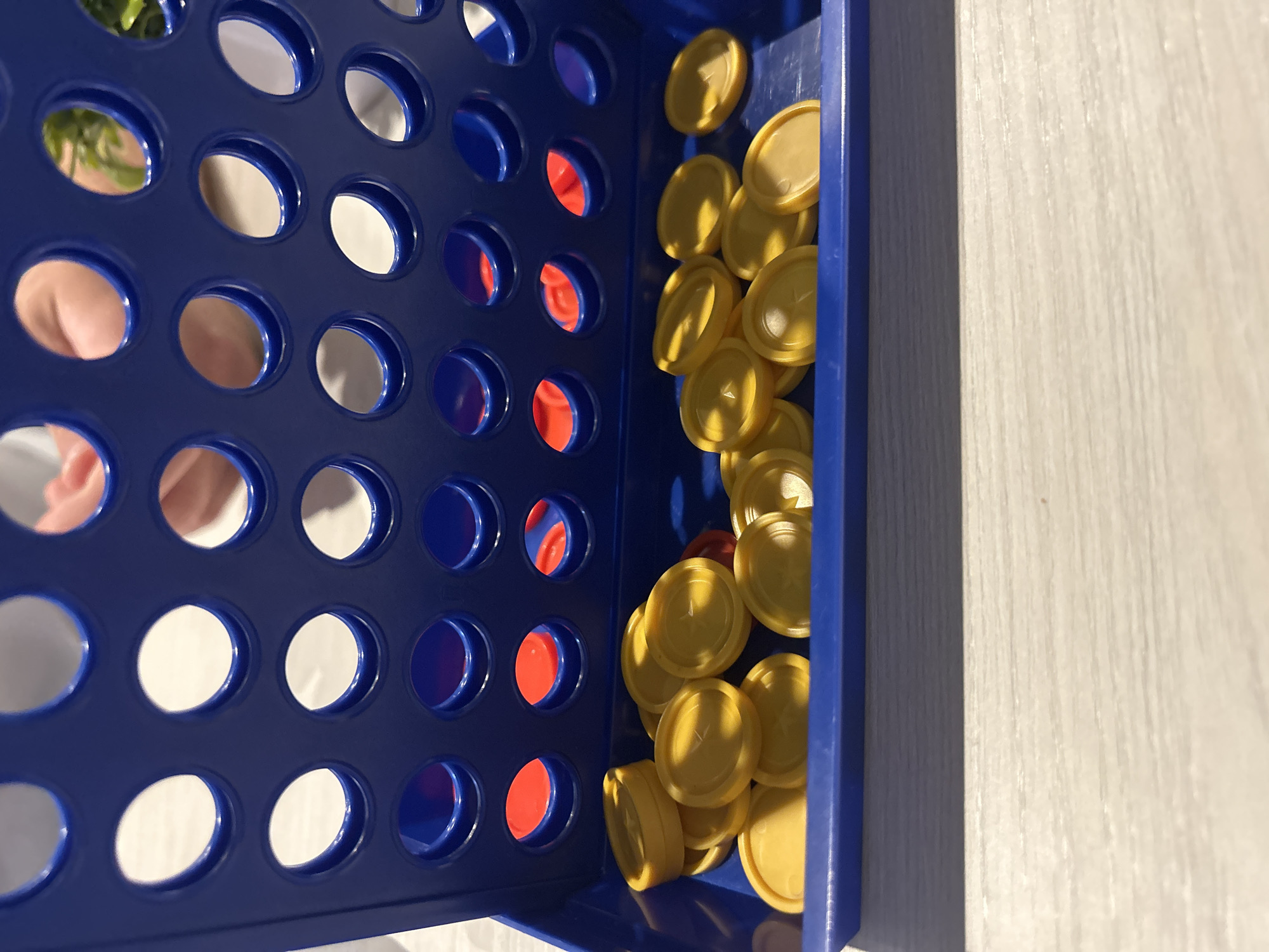 A close-up, eye-level shot shows a blue plastic Connect Four game board in a domestic setting. The game board is upright, with the slots facing the viewer. A collection of yellow circular game pieces is piled in the lower tray of the board, with a few red pieces visible through the slots in the game board. The background is blurred, with hints of hands and a green plant suggesting a residential interior. The image does not depict any elements related to the city of Chaikovsky, Russia.