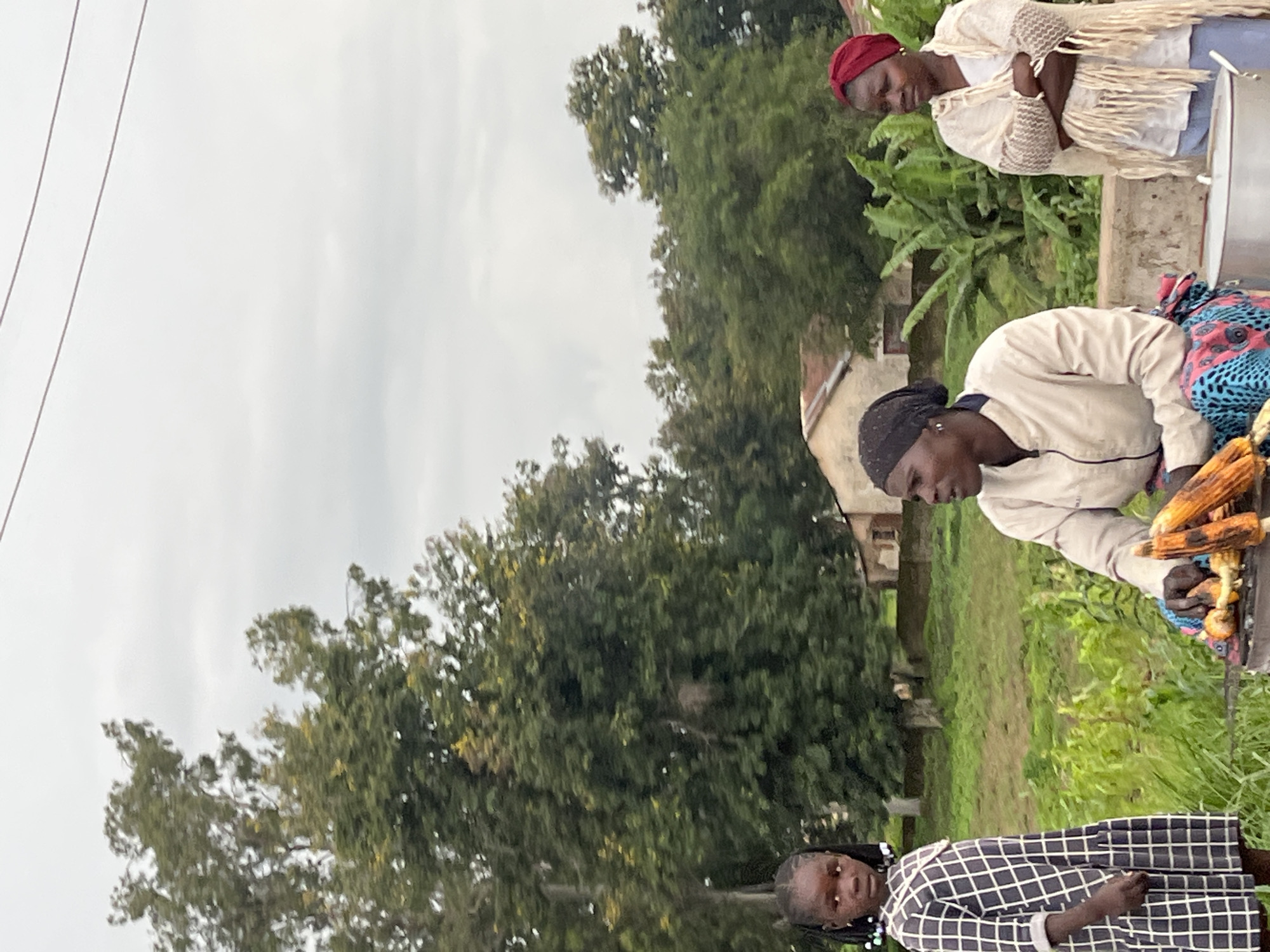 In Bukuru, Nigeria, under an overcast sky, three individuals are positioned outdoors. A young girl, standing on the left, is dressed in a dark and white checkered dress. To her right, an adult sits, wearing a light-colored jacket over a patterned skirt, actively tending to grilled corn over a small fire. On the far right, another adult stands with arms crossed, draped in a light-colored shawl with fringed edges and wearing a red headscarf. The background consists of lush green vegetation, trees, and buildings with red roofs. A large metal pot is visible in the foreground.