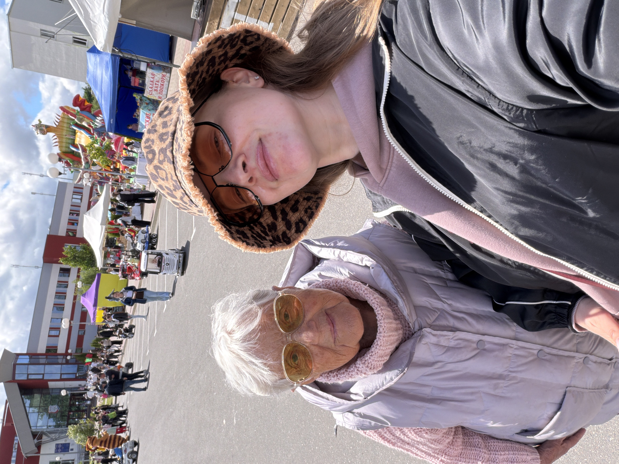 The image depicts two individuals, an older woman and a younger woman, in what appears to be an outdoor public area. The older woman has white hair and is wearing a light purple puffy jacket and glasses with round, gold-tinted lenses. The younger woman is wearing a leopard print bucket hat and round, brown-tinted glasses. The older woman is positioned in the lower part of the frame, while the younger woman is in the upper portion.
The background shows a cityscape with several tents and people. There are buildings with red and white facades, and blue and white tents are also seen in the distance. A large, colorful sculpture of a dragon-like figure is visible behind the tents. A crowd of people is milling around in the central area.
The image was taken in Valozhyn, Belarus.