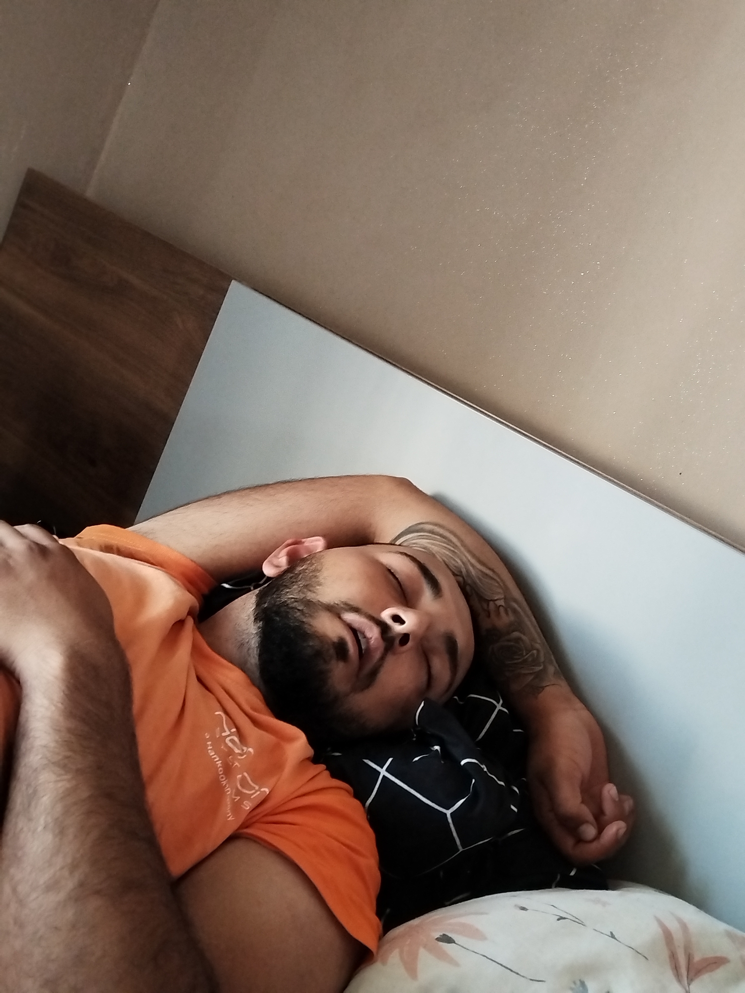 A person lies sleeping on a bed. The individual is wearing an orange T-shirt and has a tattoo on their left arm. They are positioned on a pillow with a patterned black and white pillowcase. The location is Unknown, Unknown. The environment appears to be a bedroom, suggested by the bed and pillows. The background consists of a wall with a neutral tan color and the side of a wooden dresser or headboard.
