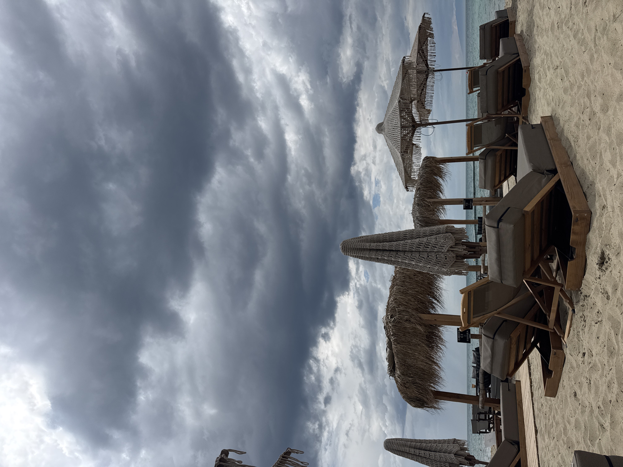 The image shows a beach scene with several thatched umbrellas and lounge chairs arranged in rows. The sky is overcast with dramatic, dark clouds covering a large portion of the frame. The chairs are made of wood with gray cushions. The beach is sandy and stretches to the horizon, where the sea and sky meet. The location is Unknown, Unknown.