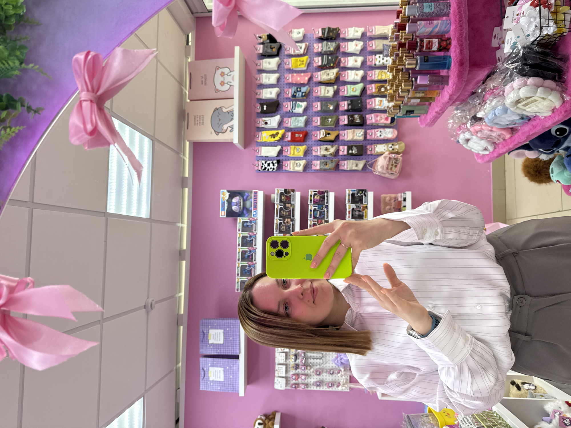 The image displays a selfie taken by a young woman in a brightly lit retail store, with the photograph oriented sideways. The woman, who has shoulder-length straight brown hair, looks directly at her phone, held up to capture the picture. She is wearing a white pinstriped long-sleeved button-down shirt and grey trousers. A silver watch adorns her left wrist, and a ring is visible on her left hand. The phone she uses is a bright neon green iPhone, equipped with a triple-lens camera system. Her right hand is slightly raised, displaying a partially open palm.
The background features a vibrant pink wall, indicating the store's decor. On the upper left, a reflection in a mirror shows a pink ribbon bow and sections of fluorescent ceiling lights. Beneath this reflection, a white shelf holds two small, rectangular items, likely notebooks or gift boxes, decorated with cartoon animal designs—a cat and an elephant. Visible text on these items includes "CAT," "ELEPHANT," and other words in an unfamiliar script.
To the right of these items, a large pegboard display is filled with numerous small accessories, possibly hair clips or keychains, showcasing diverse patterns like cow print, alongside solid colors such as white, black, and yellow. Above this, a pink tiered display shelf is stocked with many small bottles, suggesting perfumes or body sprays. Further right, another pink display contains clear plastic bags filled with pastel-colored scrunchies or hair bands in white, pink, and blue.
Below the pegboard accessories, several Funko Pop! collectible boxes are visible, with the character "Stitch" clearly identifiable on one. Plush toys, including a blue Stitch character and a brown furry animal, are also present on lower shelves. The overall setting suggests a specialty shop offering gifts, accessories, or collectibles, characterized by its colorful merchandise and bright artificial lighting.