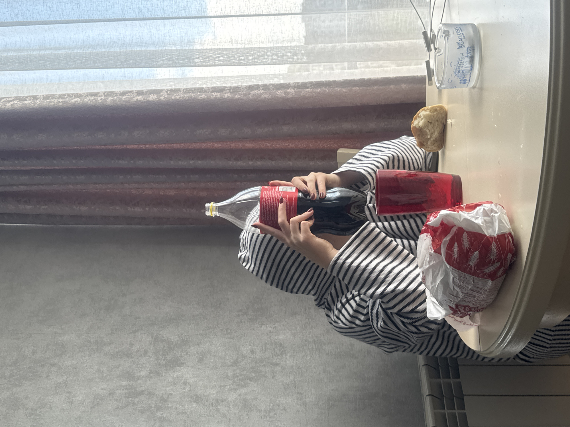 A person wearing a black and white striped garment is holding a Coca-Cola bottle and a red tumbler on a table. On the table, there is also a loaf of bread, a white plastic cup with blue and white graphics, and a plastic bag with red graphics. The table is near a window with sheer curtains, and a gray wall is visible in the background. This scene is in Tbilisi, Georgia.