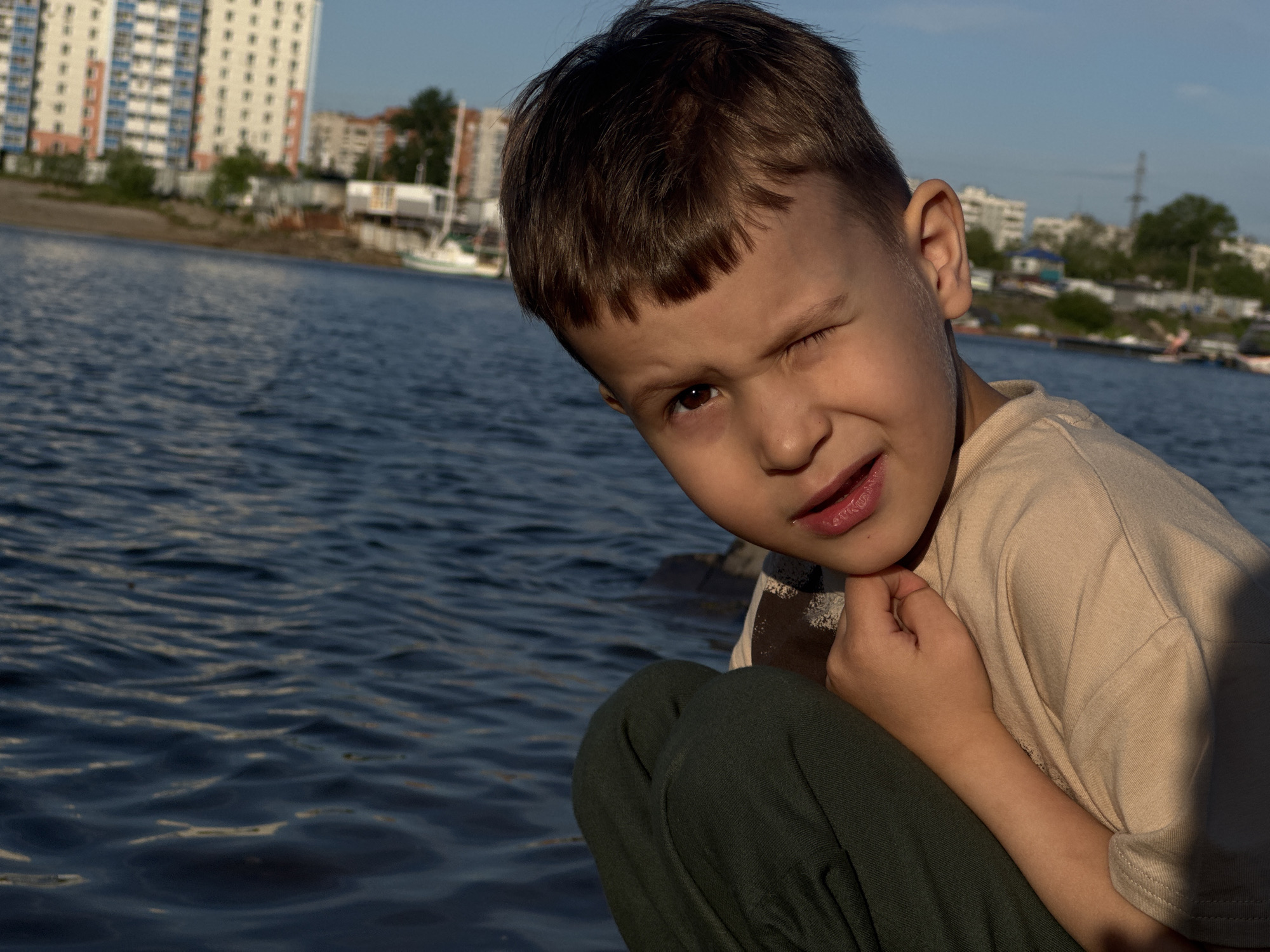 A young boy with brown hair and expressive eyes is captured in a close-up shot, winking at the camera. He is wearing a light-colored t-shirt and dark green pants, and is crouched down, resting his chin on his hand. The boy's lips are slightly parted, revealing a hint of a smile. The background features a body of water, likely a river, with gentle ripples on its surface. In the distance, along the riverbank, are several buildings, including a tall, multi-story apartment complex and smaller structures that appear to be houses or other residential buildings. A few boats are visible on the water, and some trees dot the landscape. The sky above is a clear blue, suggesting a pleasant day. The lighting indicates it is likely late afternoon or early evening, with the sun casting a warm glow on the scene. The overall mood is playful and engaging, with the boy's cheeky wink conveying a sense of fun and mischievousness. The location context of Khabarovsk, Russia, places the scene in the Russian Far East, likely near the Amur River, which flows through the city.