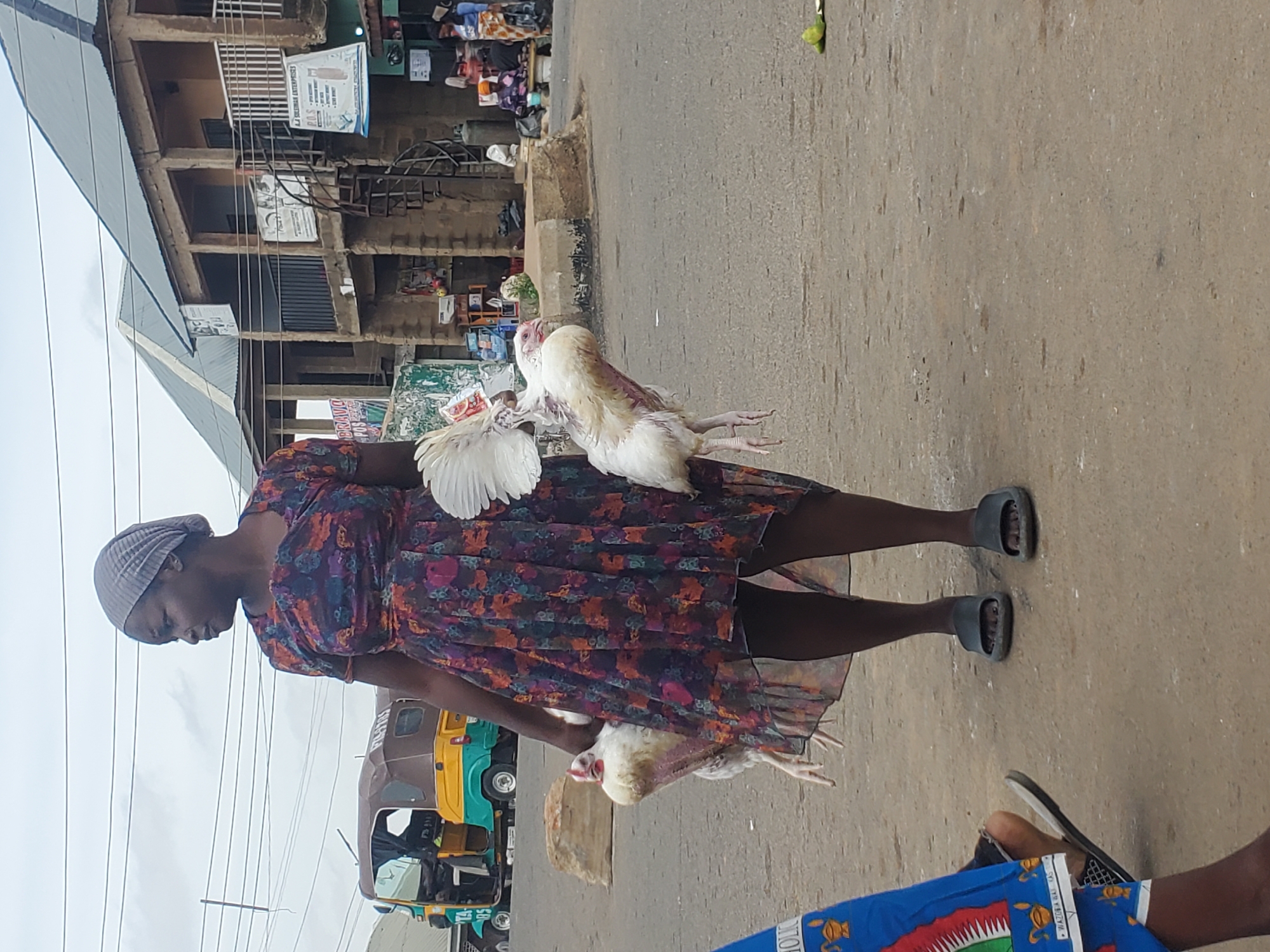 A woman, wearing a headwrap and a floral-patterned dress, is standing on a paved street in Gwong, Nigeria. She is holding two chickens. The chickens are white and appear to be dead or stunned. The woman is looking down at the chickens. In the background, there is a building with multiple stories and several shops. There is also a three-wheeled vehicle, commonly known as an "okada" or "keke napep" in Nigeria, parked on the street. The sky is overcast, suggesting it might be a cloudy day. The overall scene appears to be a marketplace or a street in a town. The woman's posture and the way she holds the chickens might suggest she is a vendor or is transporting them for a meal. There is some visible text on signs in the background, but it is difficult to read due to the image quality and orientation.
