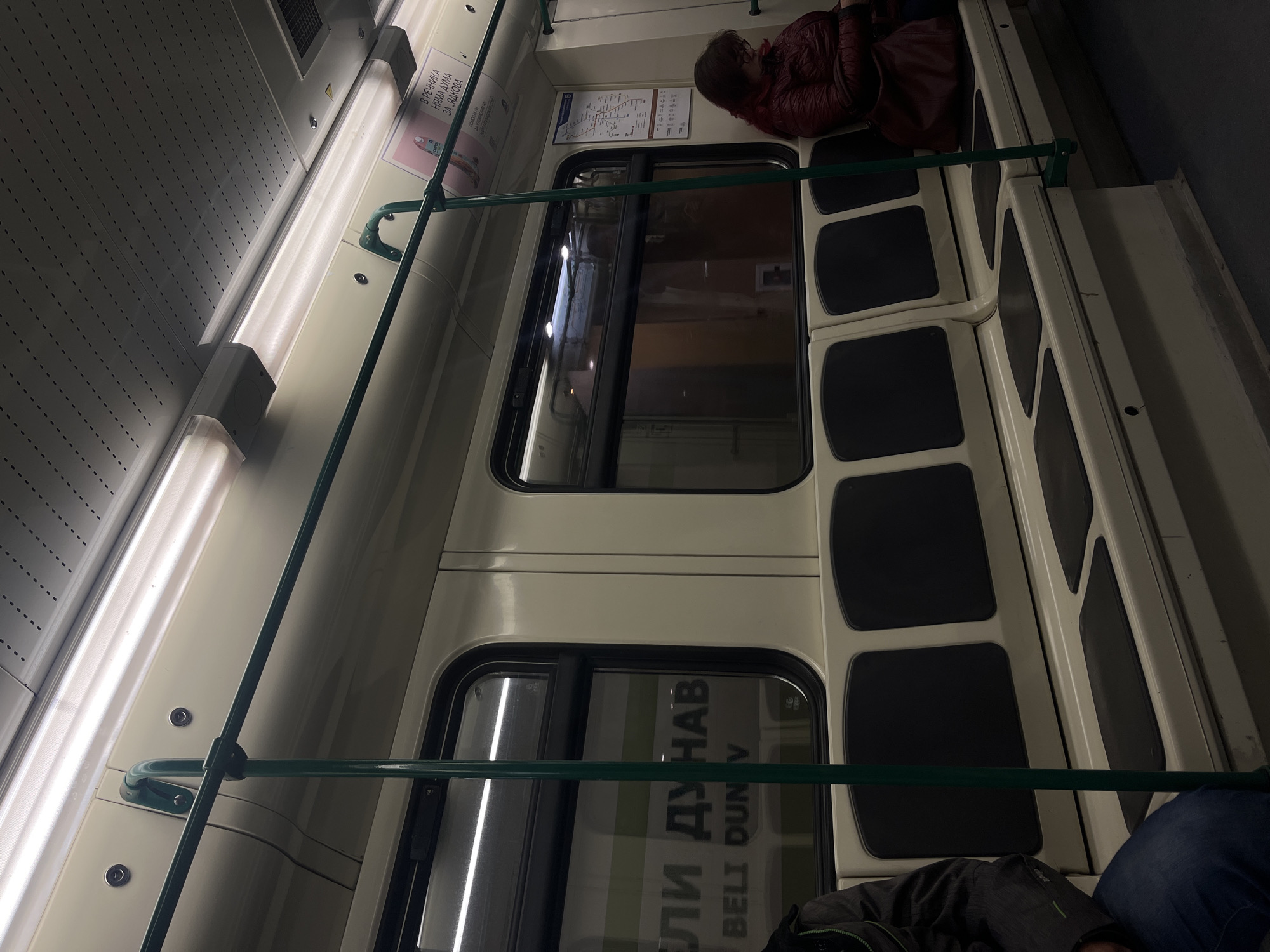 Here is a description of the image:
The photograph shows the interior of what appears to be a train or subway car. The carriage features longitudinal seating with dark padded seat cushions. Passengers are present, one seated near the top of the frame and another partially visible at the bottom. A green metal handrail runs horizontally along the car's length.
Windows are visible, one with a partial view of an exterior advertisement written in Cyrillic script, "ЕЛИ ДУНАВ BELI DUN V" . The car's interior is illuminated by a long fluorescent light panel along the top edge. Posters or signage are placed above the windows. The lighting is somewhat dim.
The image is rotated, presenting a diagonal view of the interior.
