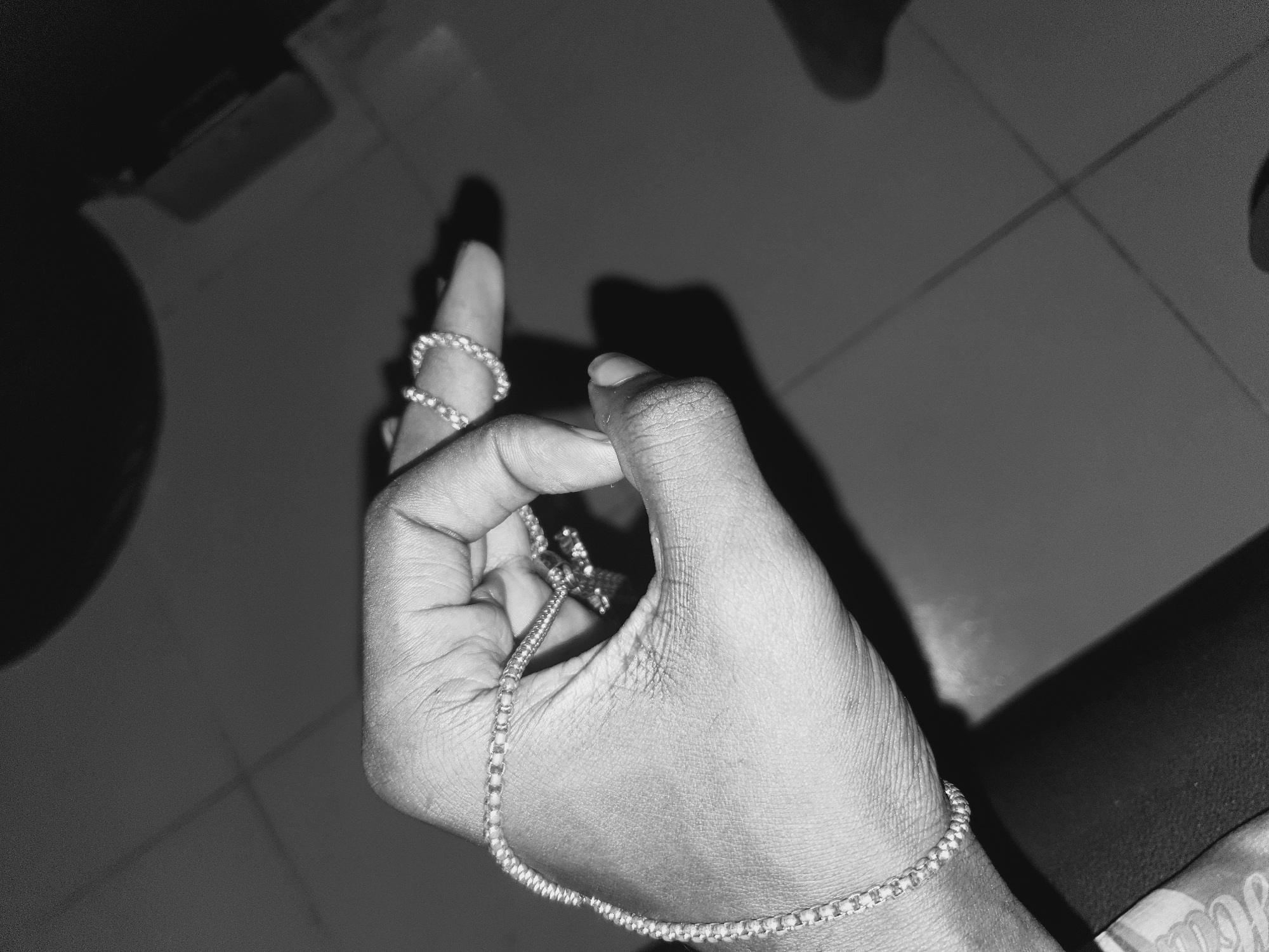 A close-up black and white image shows a person's hand adorned with jewelry. The hand is light-skinned, with visible creases and wrinkles on the skin. The index finger has a thick, beaded ring. A thin, chain-like bracelet wraps around the wrist. A longer chain bracelet runs from the wrist, between the fingers, and toward what appears to be a cross-shaped charm held loosely in the palm. The background is a mixture of tiled flooring and shadowed shapes. Based on the prompt, this could be a personal space located in Lokoja, Nigeria. The specific location cannot be identified without further context.