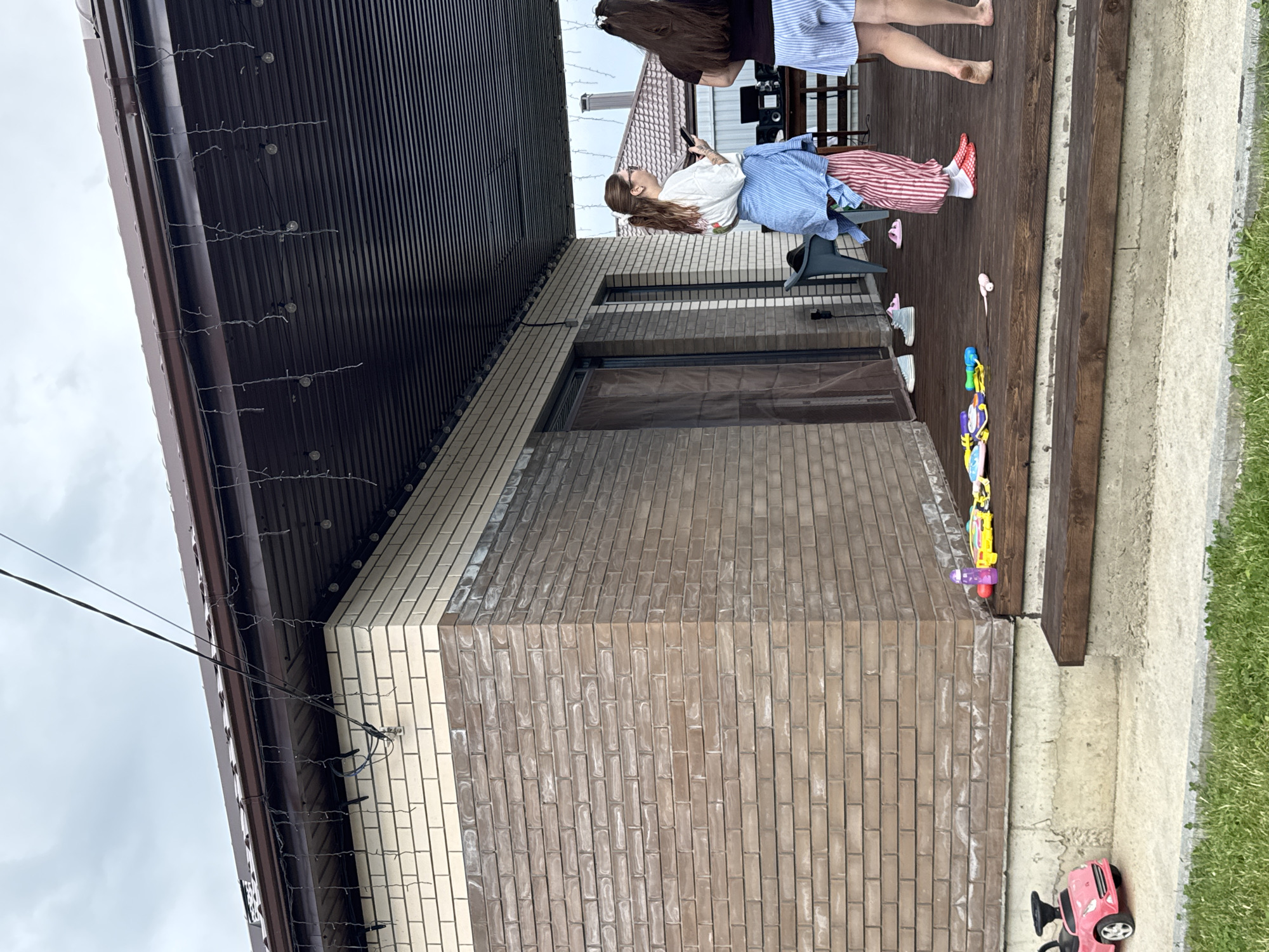 The image depicts a residential porch area outside a brick house in the village of Dyomino, Russia. Two young women are standing on the wooden deck. The woman on the left is wearing a white t-shirt, a blue open shirt, and striped red and white pants, holding a smartphone. The woman on the right is wearing a black t-shirt and blue and white striped shorts, with her back to the camera. Various toys, including a pink ride-on car, water guns, and other colorful objects, are scattered on the porch steps and deck. The porch is covered by a corrugated metal roof, decorated with strings of lights. The house has light-colored brick on the upper portion and darker brick on the lower section. A concrete foundation and grassy area are visible in the foreground. The sky is overcast.