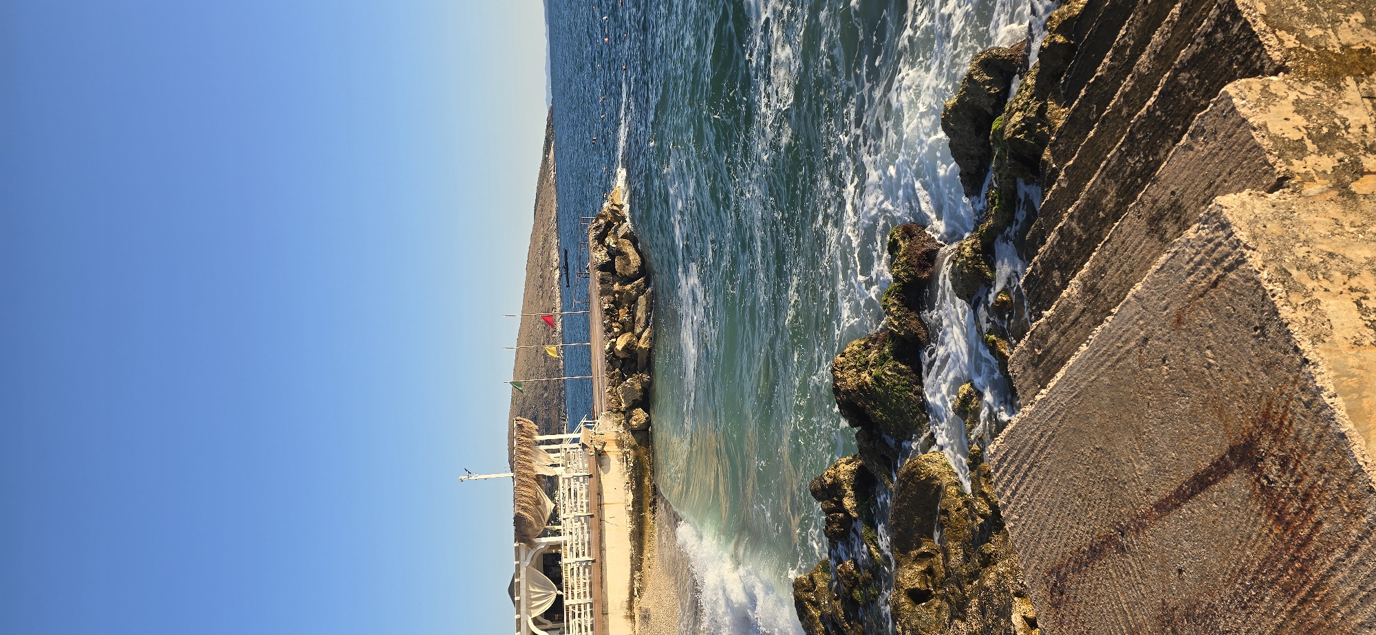 The image depicts a coastal scene in Saranda, Albania, characterized by a clear blue sky above a tranquil sea. In the foreground, rough-hewn concrete steps descend towards the water, with waves gently crashing against moss-covered rocks lining the shore. A wooden pier extends into the sea, adorned with three flags—green, yellow, and red—fluttering from poles. To the left of the pier, a white-canopied establishment with straw roofing sits on a pebble beach. The backdrop features a gently sloping, arid hill. The water is a vibrant turquoise, transitioning to a deeper blue further out, with small white-capped waves indicating moderate movement. The lighting suggests it is either early morning or late afternoon, casting warm tones on the concrete steps and highlighting the textures of the rocks and the water's surface. The scene is devoid of people, presenting a serene and natural coastal landscape.