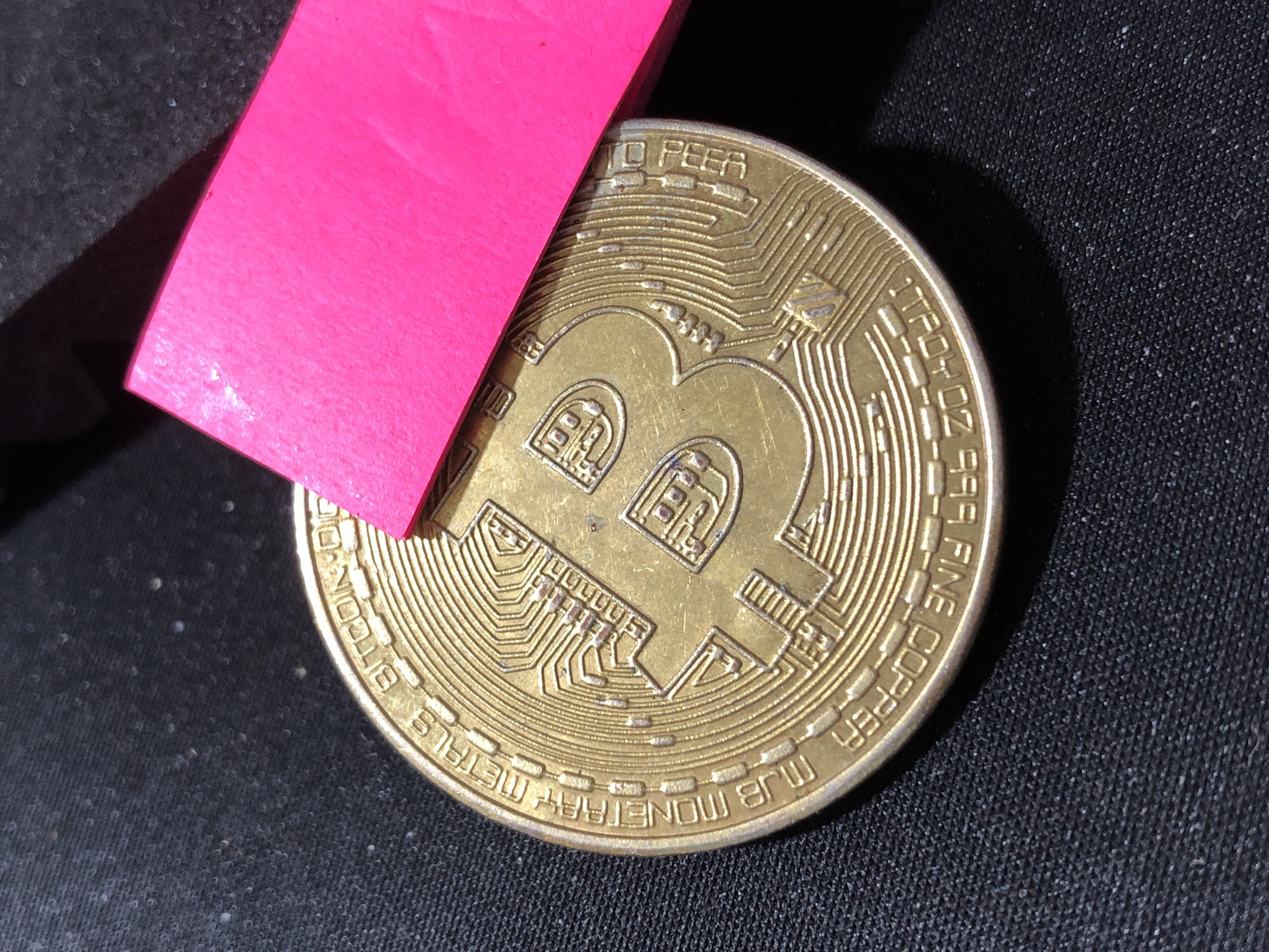 This image features a close-up, high-angle shot of a gold-colored Bitcoin coin. A bright pink rectangular object, possibly a piece of paper or plastic, partially covers the top left portion of the coin. The coin rests on a textured dark surface, which appears to be fabric, possibly a tablecloth or clothing. The lighting creates a strong shadow from the pink object onto the coin and the surface.
Visible text on the coin includes: "BITCOIN" along the bottom left curve, "TROY OZ" on the bottom right curve, and "FINE COPPER" on the lower right curve. The iconic "B" symbol, representing Bitcoin, is prominently displayed in the center of the coin. The coin has intricate circuitry-like patterns etched around the central symbol. The overall impression is one of a still life arrangement focusing on the physical representation of a cryptocurrency. There are no people visible, and the scene is static. The time of day, weather, and specific location cues are not discernible from the image itself, beyond the general context of a dark, indoor setting.
