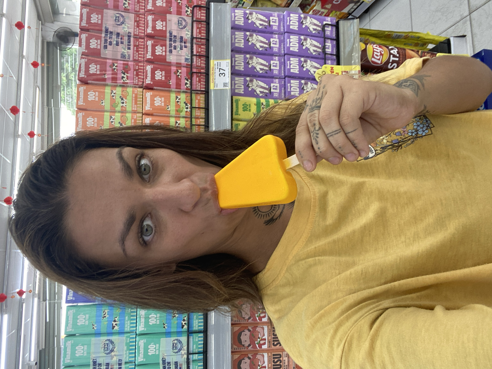 A woman is standing in the aisle of a store, possibly in Đồng Nai, Vietnam, as evidenced by the Vinamilk products. She is holding a yellow popsicle, partially in her mouth. She is wearing a yellow t-shirt and has tattoos on her arm, hand, and neck. The shelves behind her are stocked with Vinamilk and other products like "ADM" branded items and Lay's potato chips. A small fan hangs from the ceiling in the upper left corner of the image. Red decorative items are also visible. The price tag "37" is visible on the shelf.