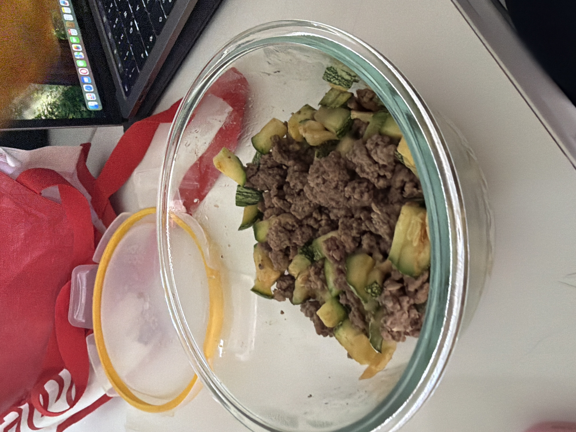 A clear glass bowl containing cooked ground meat and diced zucchini sits on a white surface near a laptop and a red bag with white accents. The bowl is positioned to the right of the laptop, and a yellow-rimmed plastic lid is beside it. The scene is on a table. This photo was likely taken indoors. Due to the lack of any context, it is not possible to confirm whether the image was taken in San Andrés Cholula, Mexico.