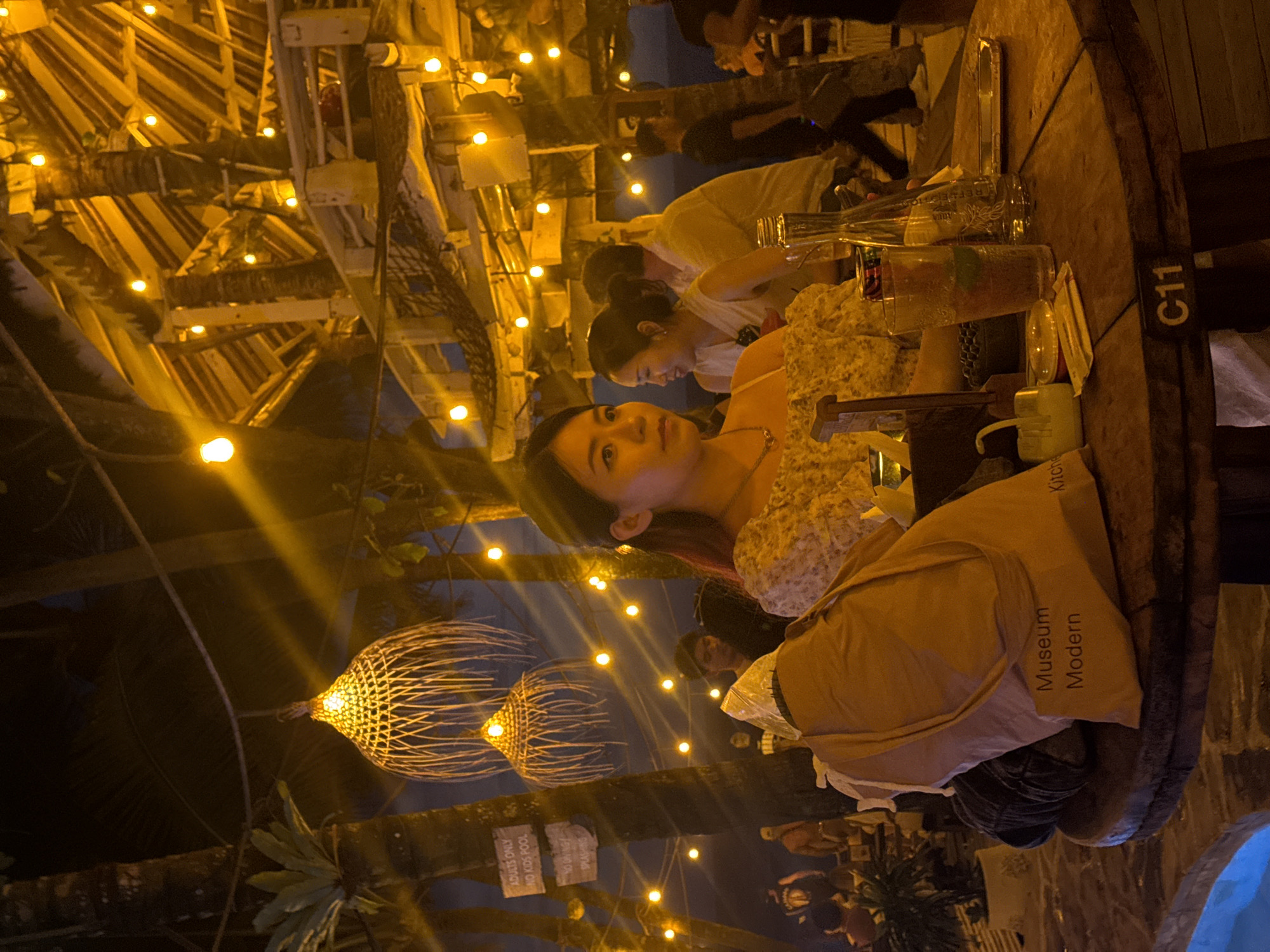 Captured in North Kuta, Indonesia, the image presents an outdoor dining scene at night. A mixed-gender group of patrons is seated at a large, round wooden table marked with "C11". On the table, there are beverages, glasses, and a bottle of "Reflections". One female subject is centrally positioned, wearing a light-colored, floral-textured top. A "Museum Modern" tote bag rests on the edge of the table. In the background, string lights and decorative woven fixtures illuminate the surrounding trees and foliage, contributing to the ambiance of the location. Signage visible among the trees reads "ADULTS ONLY NO KIDS POOL". Other patrons are discernible in the background, engaged in conversation.