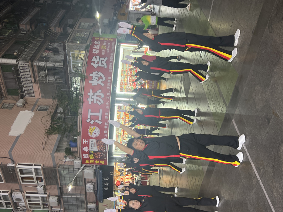 The image displays an outdoor urban scene at night in Jiangjin, China. Approximately 15 to 20 individuals, predominantly middle-aged and elderly adults, are performing synchronized exercise movements. Most participants are wearing identical black tracksuits with prominent red and yellow vertical stripes along the outer seams of the sleeves and pant legs, and white gloves. They are positioned in rows, facing generally left, with their arms extended upwards in a 'V' shape, palms directed forward. One male participant in the central foreground is directly facing the camera. The background consists of multi-story buildings, typical urban residential or commercial structures, featuring balconies and numerous external air conditioning units. A large, illuminated red sign for "江苏炒货 精品店" (Jiangsu Fried Goods Specialty Store) is prominent, with smaller text identifying the location as "江津" (Jiangjin) and a contact number (13388206667). The ground is a paved surface, and the area is brightly lit by artificial streetlights and the illumination from surrounding shopfronts.