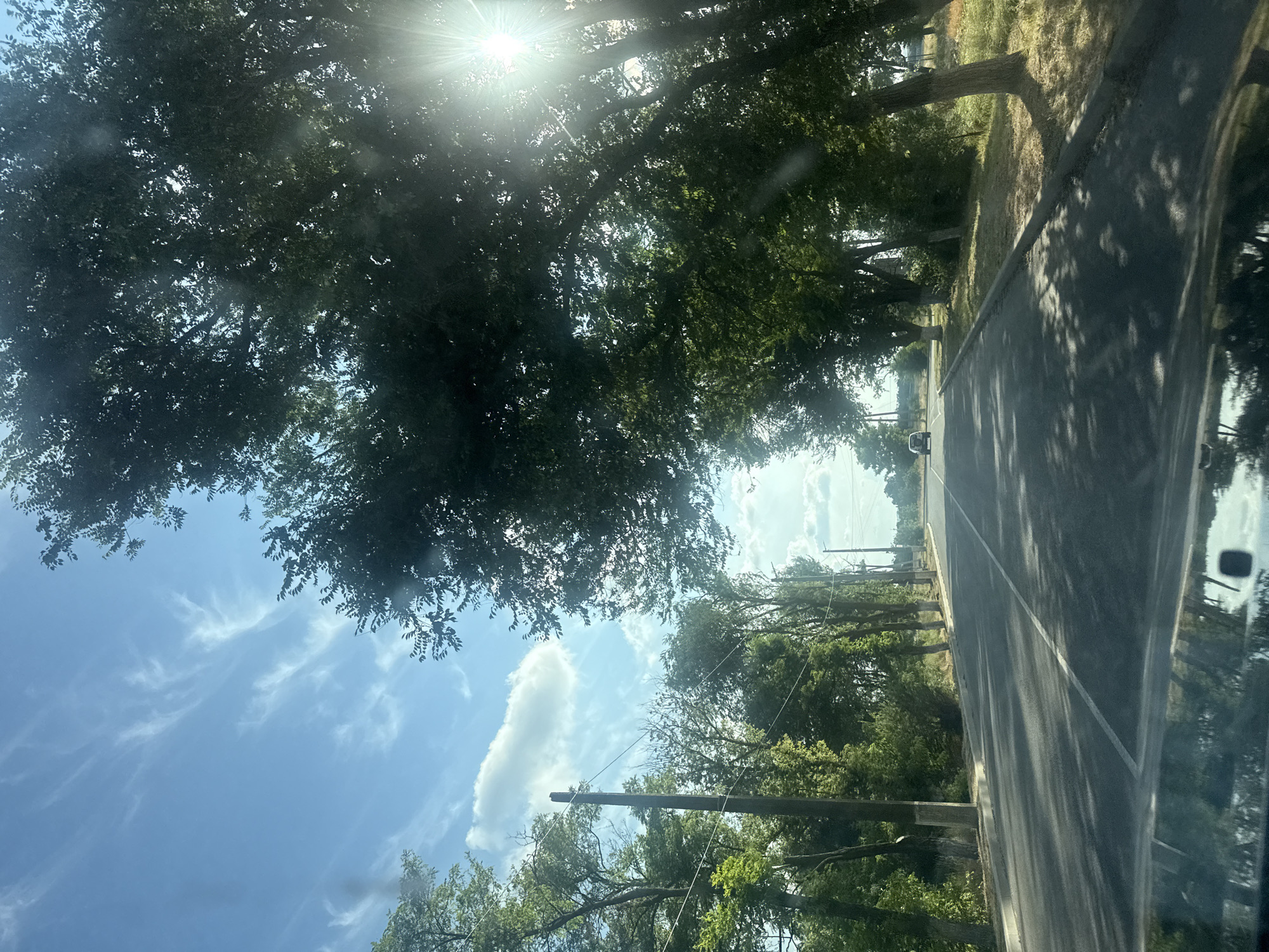 The image captures a view from a vehicle driving on a paved road lined with trees. The road has a white line marking the center and right lane. Utility poles with wires are visible along the left side of the road, interspersed with trees. The sky is blue with wispy white clouds and bright sunlight filtering through the leaves of the trees, creating dappled shadows on the road. In the distance, a dark-colored car is driving ahead on the road. The environment appears to be a semi-rural or suburban area with vegetation and a clear sky. The scene does not provide specific identifiable landmarks to pinpoint a location within Kerch, Ukraine.