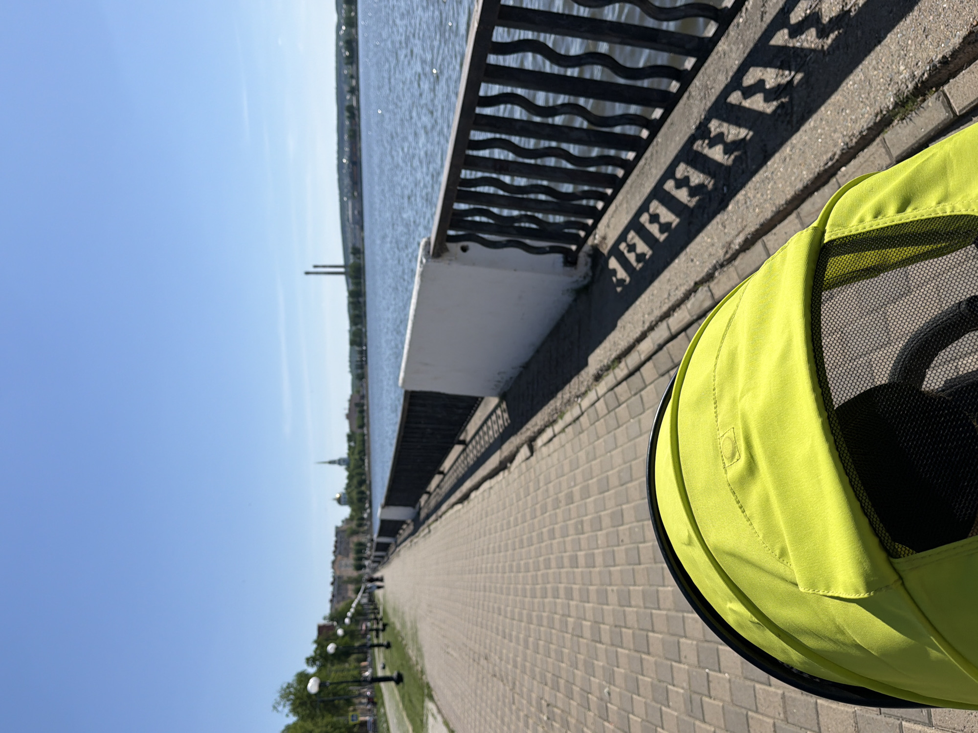 A waterfront scene in Votkinsk, Russia, shows a paved pedestrian walkway adjacent to a body of water bordered by a decorative metal railing. In the foreground, a green stroller canopy occupies the right side of the frame. In the distance, a building with a prominent steeple is visible. Along the walkway, evenly spaced street lamps line the path. The sky is clear blue.