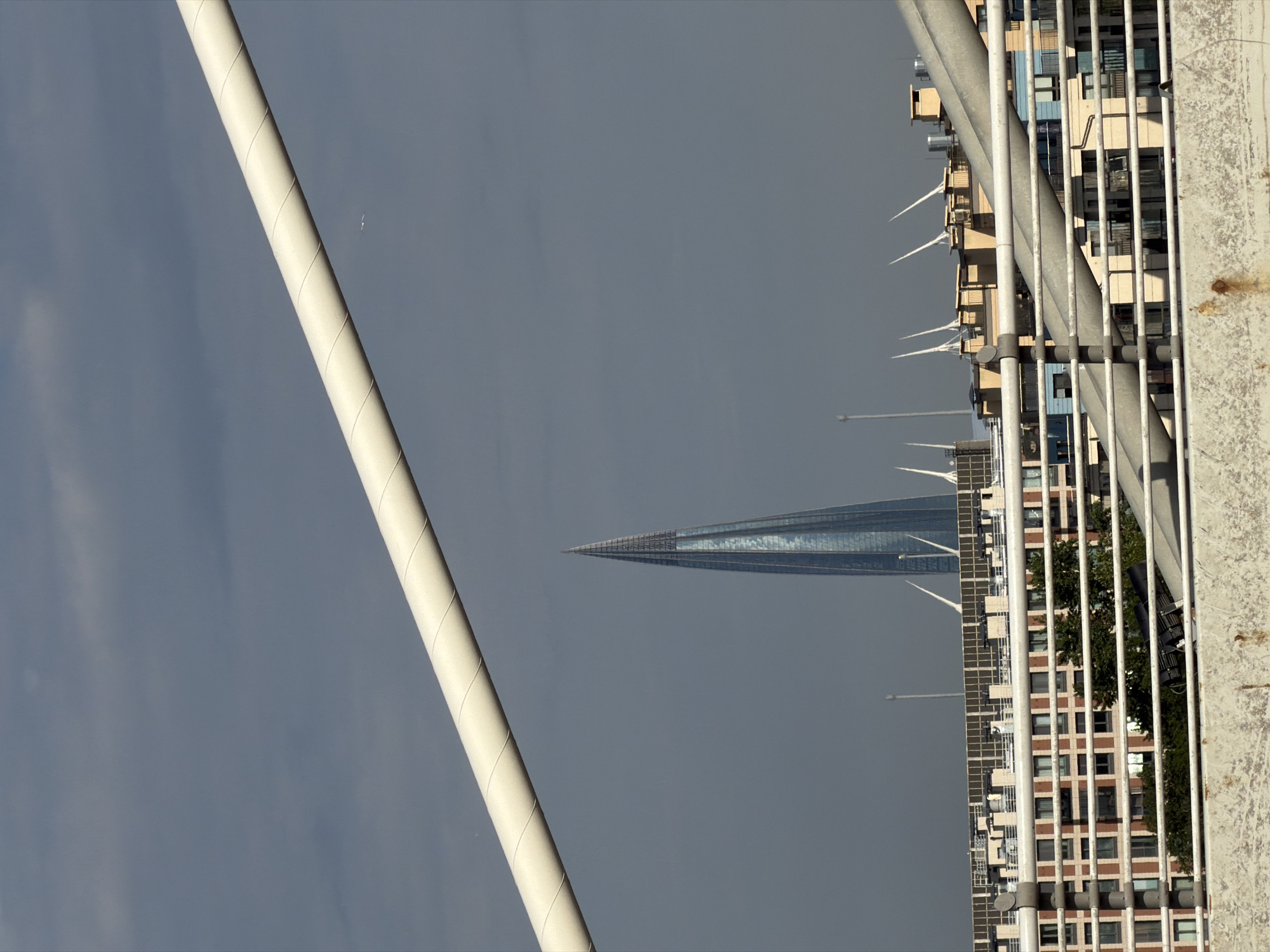 The image depicts a view of the Lakhta Center skyscraper in Saint Petersburg, Russia, seen from a distance. The skyscraper is a tall, needle-like structure with a reflective glass facade, dominating the skyline. In the foreground, there are parts of a bridge or elevated walkway, with white railings and support structures. Below the bridge, a complex of residential buildings with red brick and beige facades is visible. The sky is overcast with dark gray clouds. The overall scene suggests an urban environment with modern architecture and infrastructure.