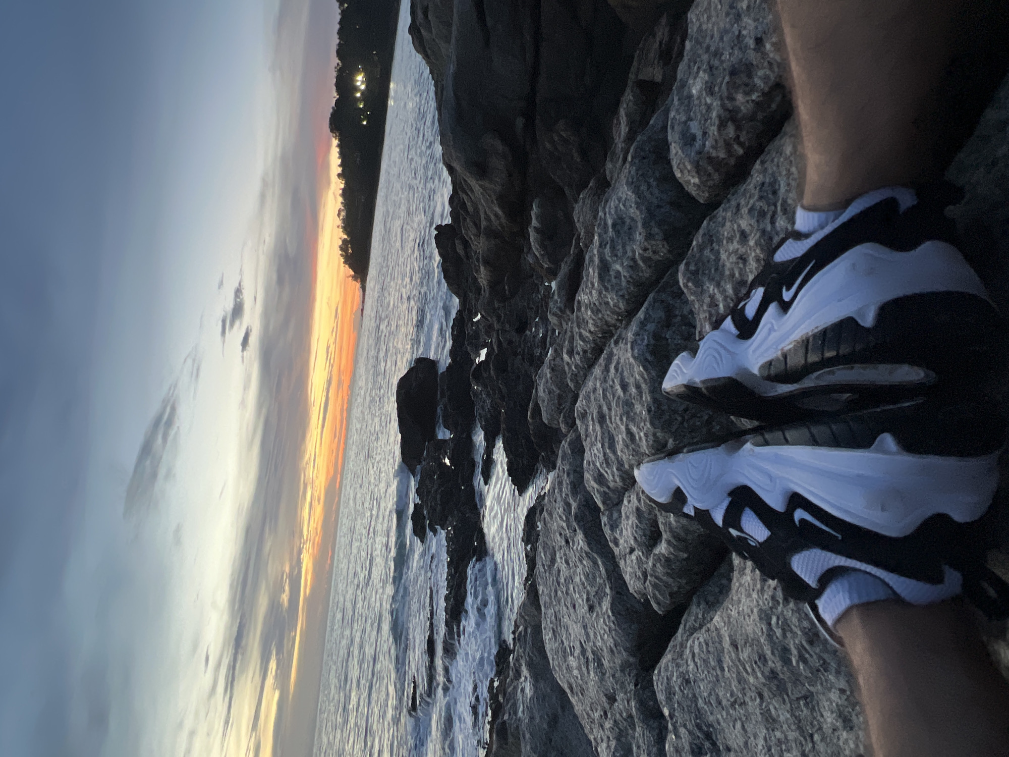 The image shows a first-person perspective of a person wearing black and white athletic shoes, sitting on a rocky shoreline at dusk. The sky displays a gradient of colors, from deep blue at the top to orange and yellow near the horizon, indicating sunset. The sea is calm with small waves lapping against the rocks. In the distance, a tree-lined coastline is visible with some lights from buildings or streetlights. The overall scene evokes a sense of tranquility and natural beauty.