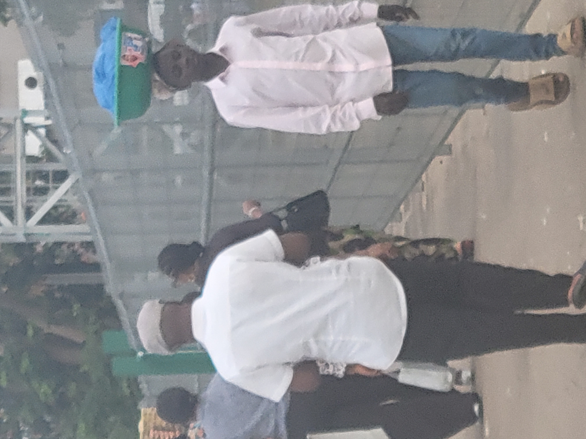 The image shows a street scene with several people in Abuja, Nigeria. A young man in a white shirt and blue jeans is balancing a blue and green object on his head. He is walking on a paved sidewalk, near a metal structure. Nearby, two people are wearing white and light blue, respectively. Another person is dressed in black. The environment appears to be urban, with a sidewalk and some greenery visible.
