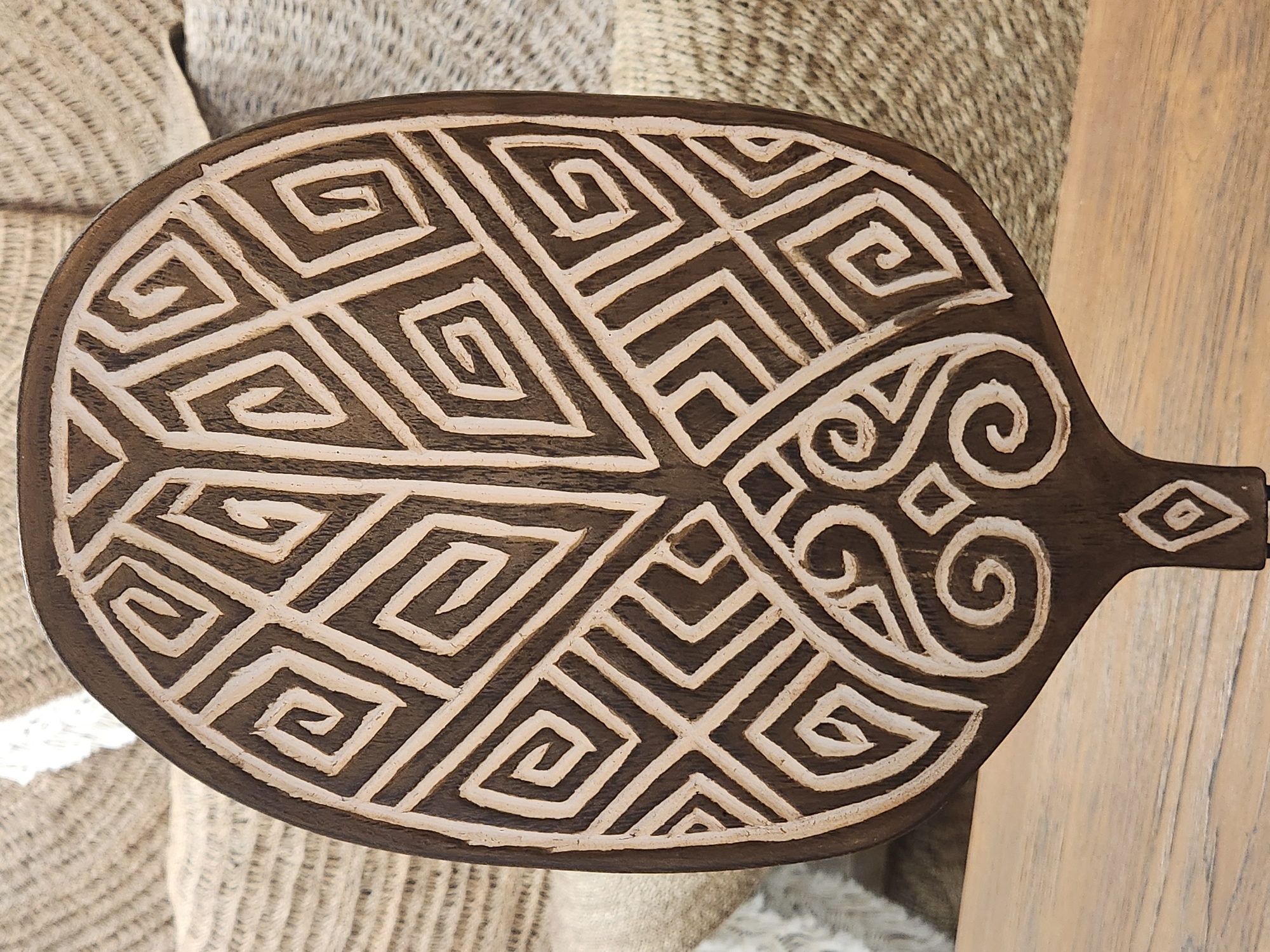 An ornate, dark brown, oval wooden object with a carved, spiraling geometric pattern filled with a light color stands upright on a light brown wooden surface. The object is likely a decorative piece or a tool, possibly a ceremonial paddle or serving board, given its shape and detailed carving. In the background, out of focus, are woven materials in neutral tones, suggesting it is displayed in an indoor setting, possibly a shop or a home. The lighting is soft, illuminating the carved details of the object. The image does not contain any discernible people, actions, or interactions. The specific location of Maitland, United States of America, is not identifiable from the visual information present in the image.