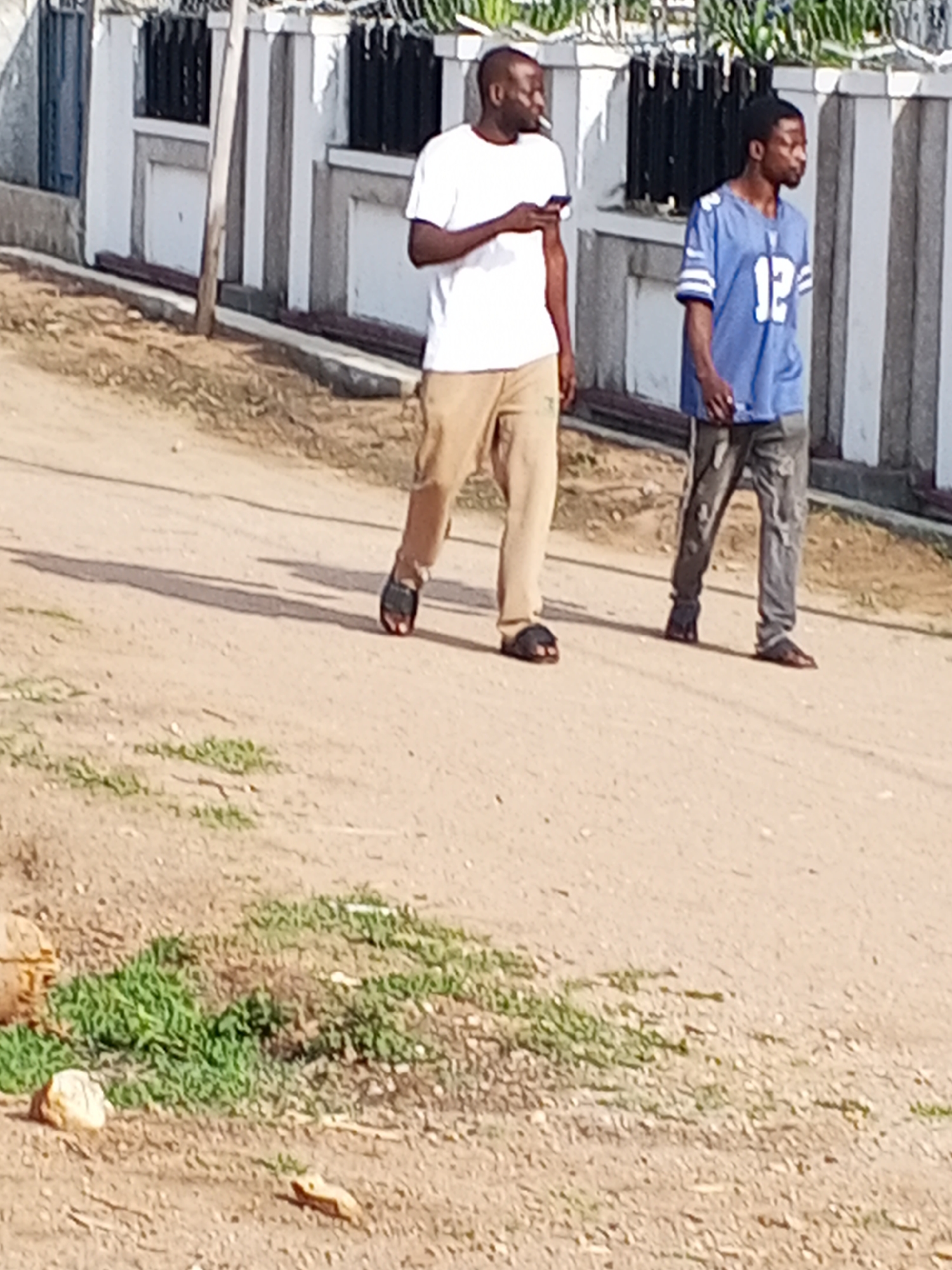 Two men are walking on a dirt path in what appears to be Adonkolo, Nigeria, under clear, sunny daytime conditions.
The man on the left has dark skin and short dark hair. He is wearing a plain white short-sleeved t-shirt, light brown or khaki pants, and dark flip-flops. He is holding a white smartphone in his right hand and has a light-colored object, possibly a cigarette or gum, in his mouth. He is looking towards the right side of the frame.
The man on the right, also with dark skin and short dark hair, is wearing a blue short-sleeved sports jersey with the number "12" prominently displayed in white on the front. He has on dark, possibly distressed, long pants and dark slip-on shoes or sandals. He is looking slightly ahead and to the right.
Behind the men, there is a low wall or fence made of concrete blocks, painted with alternating white and gray or black vertical stripes. A dark metal fence runs along the top of this wall, and some sparse green foliage is visible beyond it. The ground is a dry, light brown dirt path with patches of green grass and scattered small rocks. Shadows cast by the men suggest the sun is fairly low in the sky, indicating morning or late afternoon.