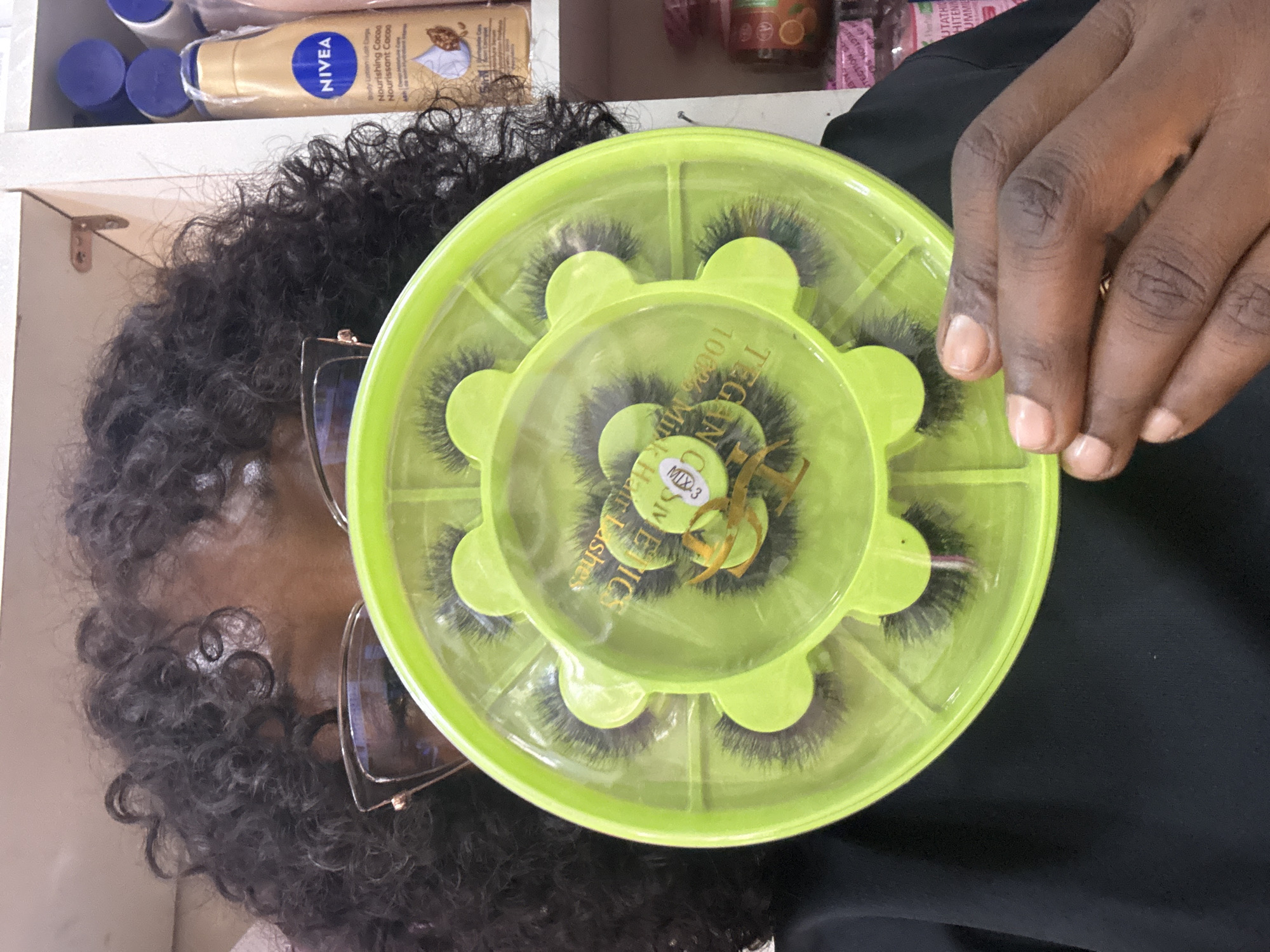 A person with dark, curly hair and glasses is holding a bright green plastic case containing multiple pairs of false eyelashes. The case is circular with compartments for each pair of lashes. The person's hands are visible, holding the case. In the background, shelves are stocked with various beauty products, including a bottle of NIVEA body lotion. The scene appears to be a retail environment, likely a beauty store or salon. The image is likely from Jos, Nigeria, given the user's prompt.