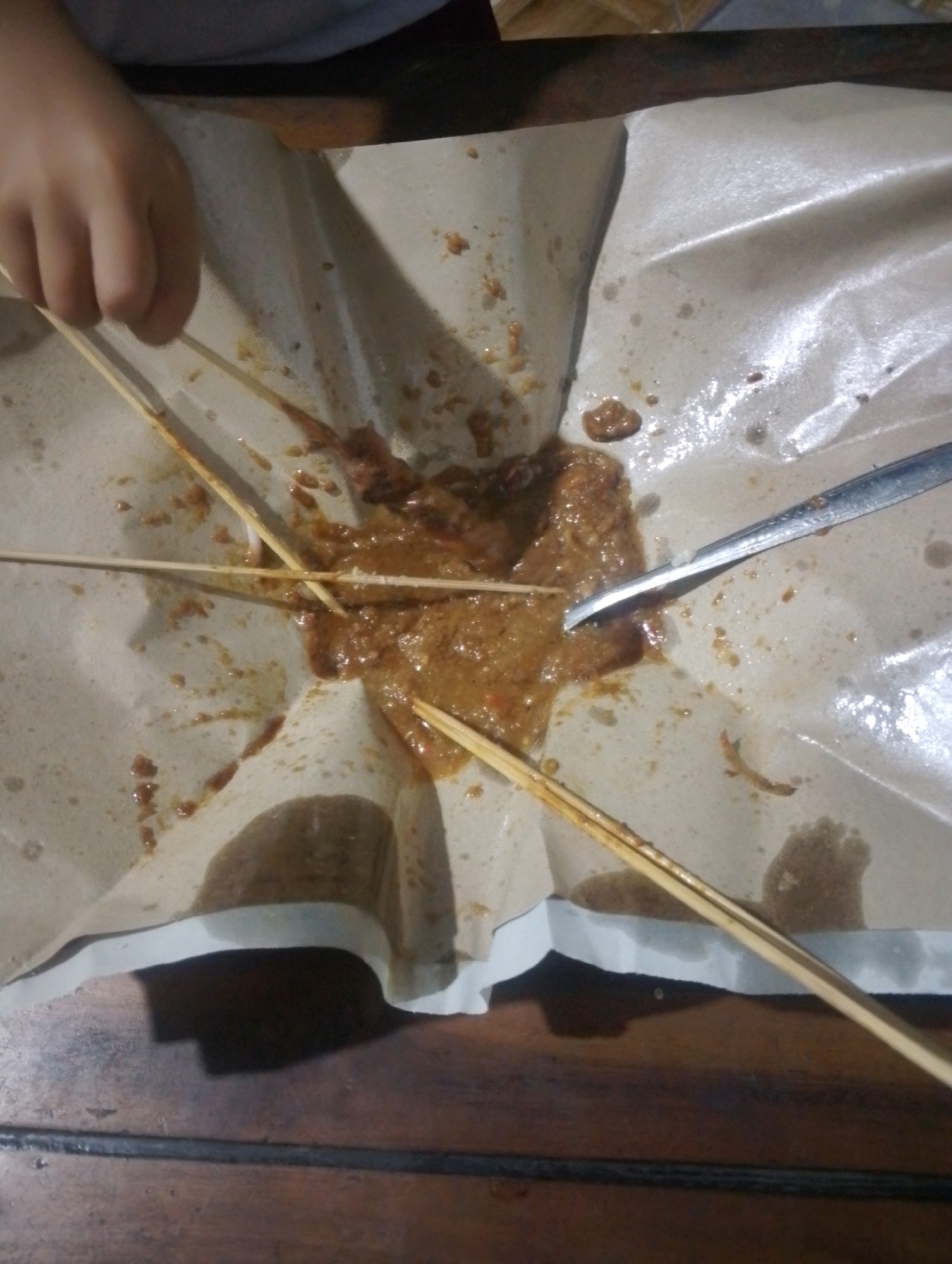An overhead shot shows a messy spread of food on a brown paper covering a wooden table. The food appears to be satay, as evidenced by the skewers and the peanut sauce that has splattered across the paper. A person's hand, with fingers curled, is visible in the upper left corner, appearing to reach for a skewer. A metal spoon rests near the right edge of the frame, also covered in sauce. The scene suggests a casual meal, possibly outdoors, given the rustic setting and the way the food is presented. The location is identified as Kendalrejo, Indonesia.