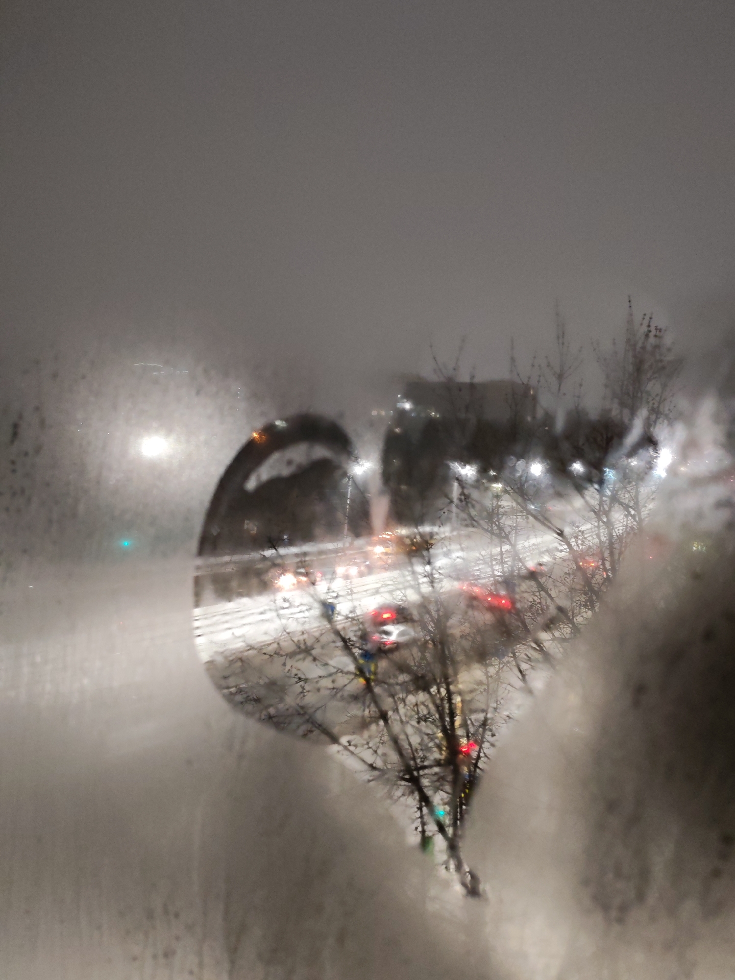The image shows a blurred view of a snowy street in Izhevsk, Russia, through a fogged-up window. Cars with their headlights on are visible on the road, and bare trees line the street. The scene appears to be at night.