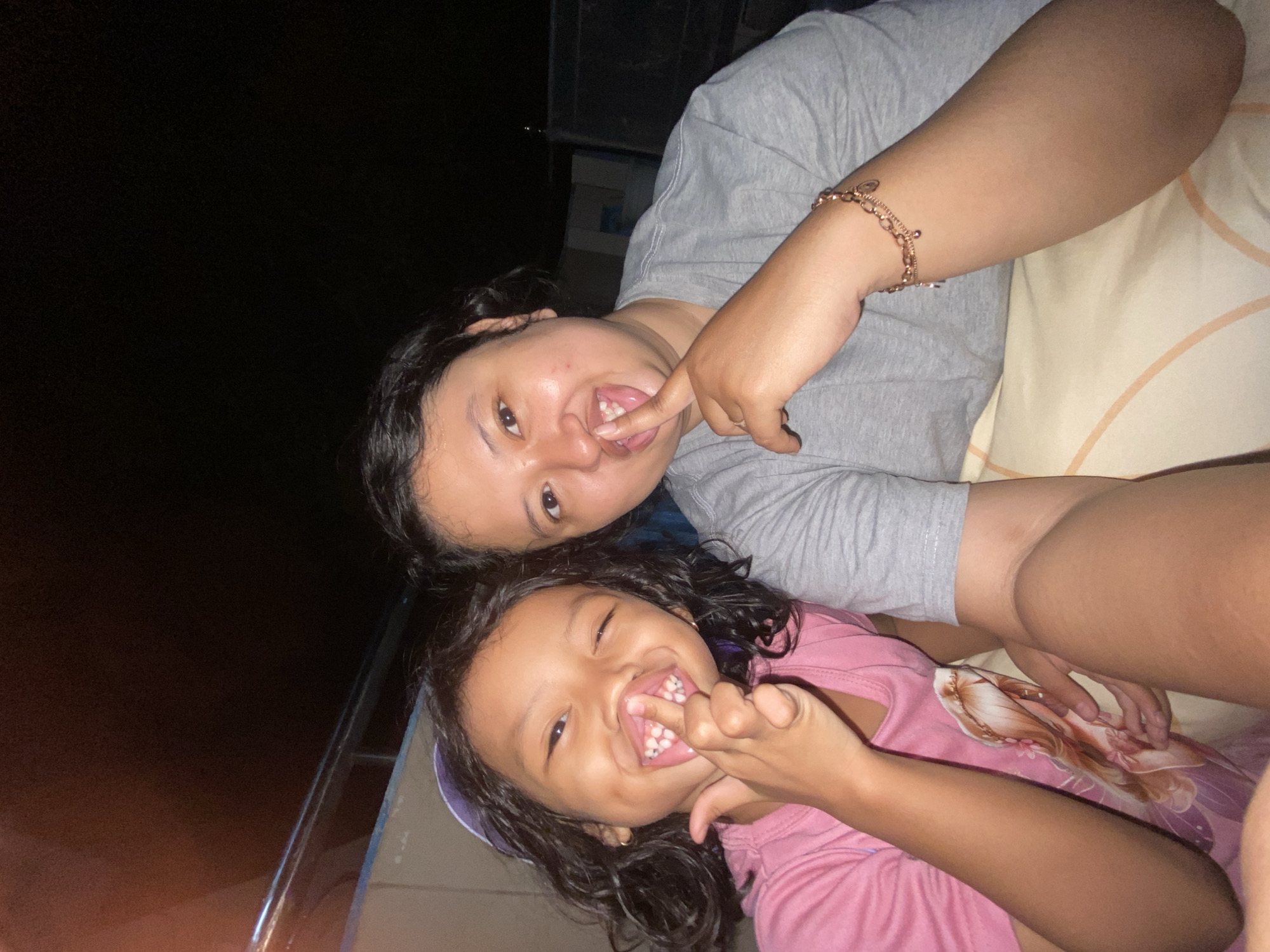 In Gelam, Indonesia, an image captures two individuals, an adult and a child, posing for a selfie outdoors at night. The adult, wearing a grey t-shirt, has their right index finger raised to their lips, with a bracelet adorned with charms on their left wrist. The child, in a pink shirt, also has their right index finger near their mouth, and both subjects display a playful expression with their teeth visible. The background is dark, suggesting an evening setting, with a hint of a railing and a dimly lit structure.