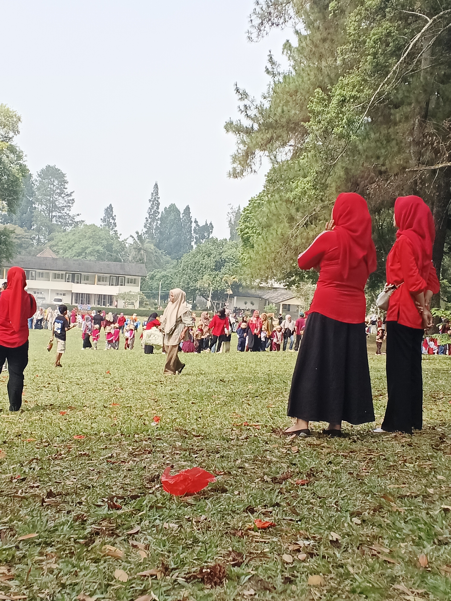 The image depicts an outdoor event on a grassy field, likely in Sudajaya Girang, Indonesia, with a building and trees in the background. In the foreground, two individuals wearing red hijabs and black skirts are standing, facing away from the camera. To the left, another person in a red hijab and black pants is also visible. The middle ground is populated with a group of people, including children, some of whom are wearing hijabs. Some individuals appear to be engaged in an activity or observing something. The environment is characterized by a large grassy area with scattered leaves and some fallen red petals. The overall atmosphere suggests a community gathering or event.