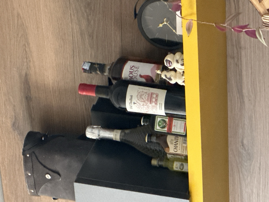 The image displays a cluttered shelf, likely within a residence, against a light-brown wood grain wall. The shelf itself is a prominent bright yellow.
From left to right, essential objects on the shelf include:
- A large, dark rectangular object, possibly a box or speaker, partially visible on the far left.
- Behind smaller items, a tall, slender bottle with a silver foil-wrapped cork, indicative of sparkling wine or champagne.
- In front of this, four miniature liquor bottles are lined up: a small clear bottle with light amber liquid, a "CHANTH" branded bottle with brown liquid, a green "Jägermeister" bottle featuring its deer logo, and another small bottle with dark liquid.
- A standard-sized dark red wine bottle with a red foil cap. Its label clearly reads "Old Tbilisi" and depicts an illustration of the iconic Ferris wheel and structures found at Mtatsminda Park Funicular in Tbilisi, Georgia, confirming the geographical context.
- Next to the wine bottle, a partially obscured amber-liquid bottle has a label featuring a prominent red rooster, with visible text "FAMOUS HOUSE".
- Two small, cream-colored, stylized figurines are positioned in front of the bottles: one with a serious expression and horn-like protrusions, and another with a wide, open-mouthed smile.
- On the far right, a round, black analog clock with golden hour and minute hands is visible.
- A plant stem with several reddish-purple leaves cascades downwards from the bottom right of the frame.
Above the yellow shelf, a portion of a dark, possibly leather, object with silver-colored studs and a flap closure is visible against the wooden wall. No people are present, and no actions or interactions are depicted beyond the static arrangement of objects. The environment suggests an indoor setting.