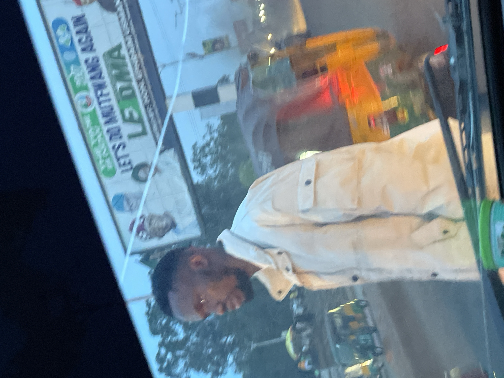 The image shows a man in a light-colored jacket standing in what appears to be a vehicle, with a blurred background of a city street. Above and behind the man is a billboard with political campaign messages. The billboard features text that reads "LET'S DO MUTFWANG AGAIN" and "LE DIMA". There are images of people on the billboard, suggesting it is a political advertisement. In the background, there are other vehicles, including what looks like a yellow and green auto-rickshaw, and trees. The lighting suggests it is daytime, possibly late afternoon or early evening, with some of the lights from vehicles and street lamps beginning to appear. The overall scene is one of urban traffic and political campaigning in Jos, Nigeria.