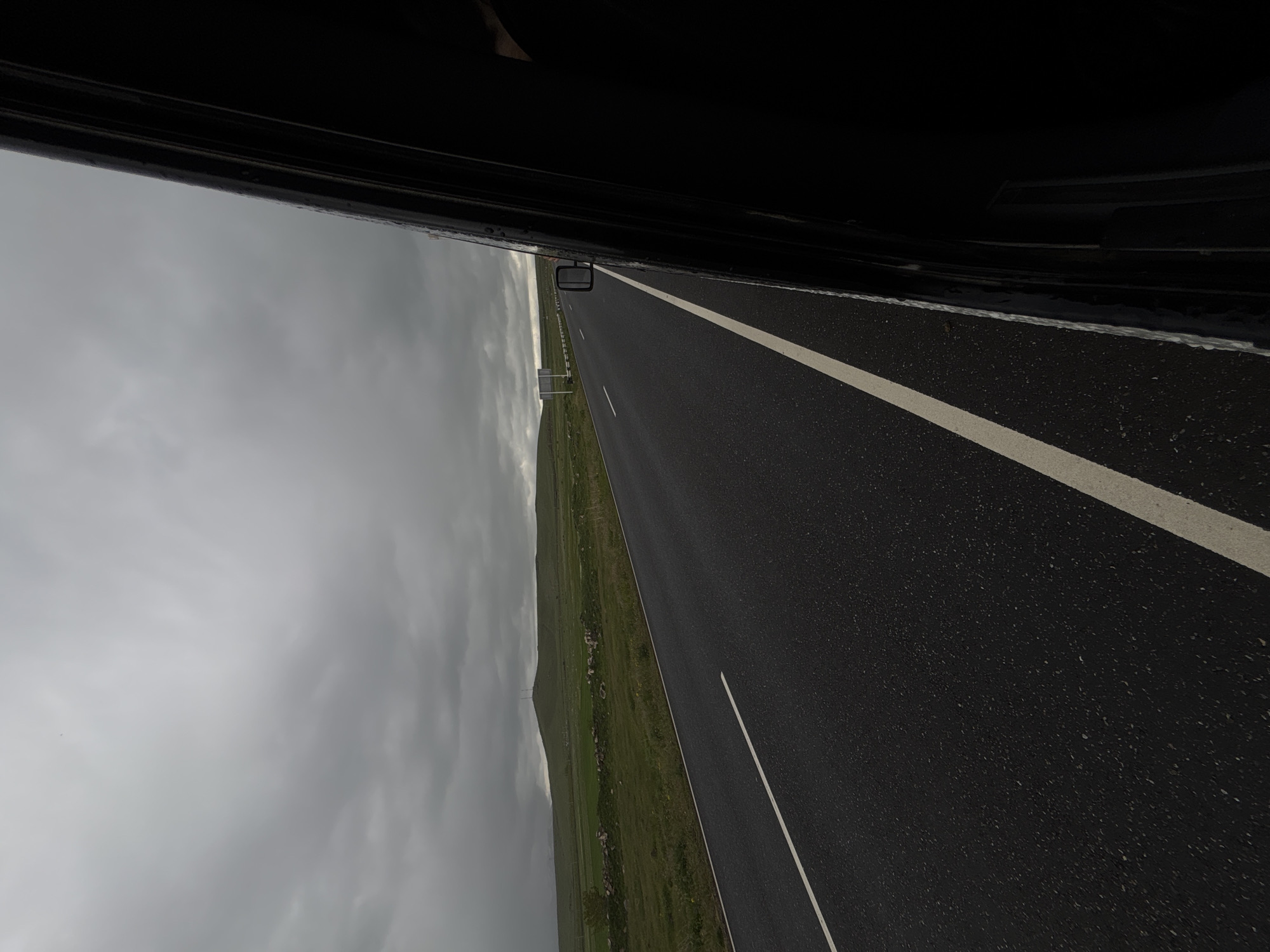 The image shows a view from inside a vehicle, likely a car, traveling on a road in Tlik, Armenia. The road is paved with dark asphalt and has painted white lane markings. To the left of the road is a grassy field with a hilly terrain. Above the field is a gray, overcast sky. Parts of the vehicle, including the frame and side mirror, are visible at the top of the image.