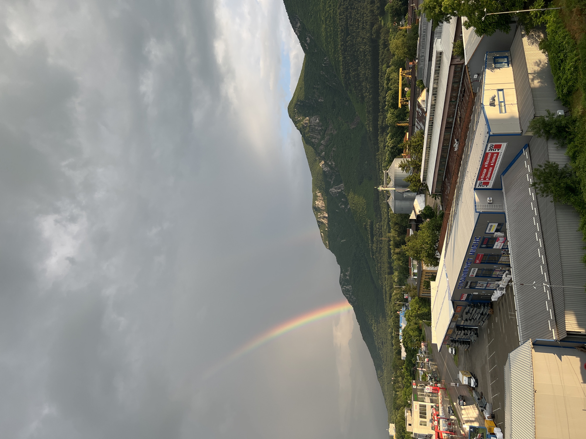 This high-angle, outdoor photograph captures a suburban/industrial landscape at the base of mountains in Vratsa, Bulgaria, under a dramatic sky featuring a double rainbow.
The sky occupies the top third of the image, dominated by heavy, dark grey storm clouds with patches of lighter grey and white where sunlight breaks through. A prominent, vibrant primary rainbow arches from the left side of the frame, touching down behind a low building towards the center-left. A fainter, secondary rainbow is visible just outside and above the primary one, particularly on the right side of the arc, above the mountain peak. The lighting suggests late afternoon or early evening, with a mix of overcast conditions and direct sunlight illuminating parts of the landscape.
The middle ground is dominated by a range of verdant, tree-covered mountains, likely the Vratsa Balkan. The mountains feature steep slopes, rocky outcrops, and dense green forests, with some peaks catching direct sunlight, highlighting their contours.
The foreground and lower-midground reveal an urban environment characterized by commercial and industrial structures. On the left, a multi-story white building with red accents, possibly a gas station or car service center, is visible with several vehicles parked nearby. Adjacent to this, a large commercial building identified by a sign that reads "СТРОЙМАРКЕТ ИНЖЕКС" (Stroimarket Injex) has multiple storefronts displaying logos such as Makita, Bosch, Gardena, Karcher, and Stihl. This building's parking lot contains a white delivery van and several cars. Further to the right, behind this building and closer to the mountains, are several large, silvery industrial silos (likely grain elevators) and other corrugated-roofed industrial buildings. A yellow gantry crane is visible in the background, signifying industrial activity. Another commercial building with a sign displaying "BAU Фасади Мазилки Замазки Лепила" (BAU Facades Plasters Putties Adhesives) is partially illuminated by the setting sun, casting long shadows. Numerous trees and patches of greenery are interspersed among the buildings. No specific individuals are discernible in the image.