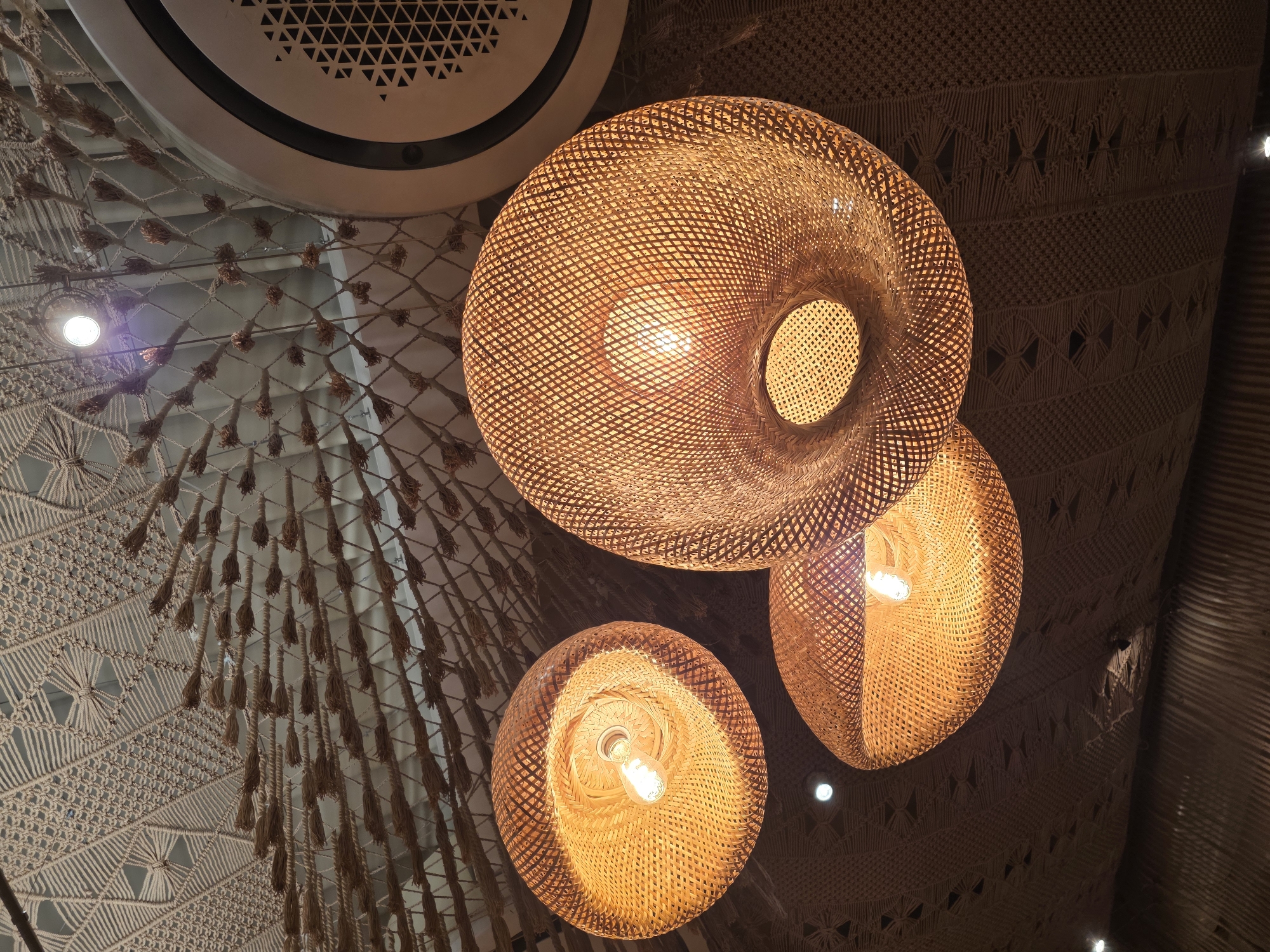 The image shows a close-up, low-angle shot of three woven pendant lamps hanging from a macrame-decorated ceiling. The lamps are made of a light-colored, natural material, likely bamboo or rattan, and emit a warm, yellow light. The ceiling is adorned with intricate macrame patterns and rows of small, tasseled embellishments. In the upper right corner, a circular air conditioning vent is visible. The overall aesthetic suggests a bohemian or naturalistic interior design. There are no people or animals present in the image. The image was taken in Varna, Bulgaria, at a restaurant called "The Cove."
