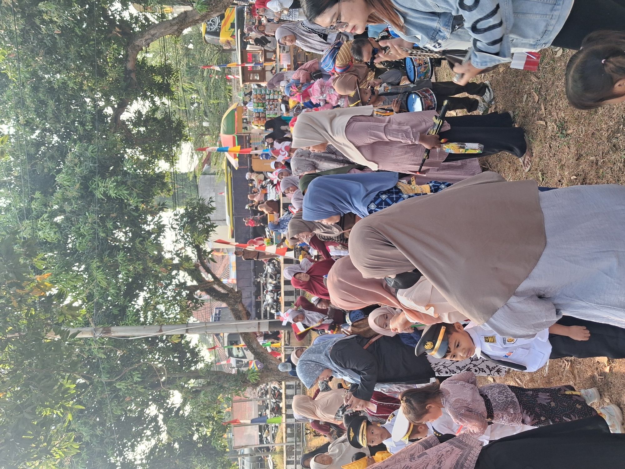 The image depicts an outdoor gathering of people in Balapulang, Indonesia. The scene appears to be a public event, possibly a festival or parade, given the presence of children in costumes, musical instruments, and decorative flags.
In the foreground, several individuals are visible. A young boy, dressed in a white uniform with a captain's hat, stands attentively. To his left, a young girl wears a traditional Indonesian outfit, a kebayak, and a batik skirt. Their attire suggests a cultural or celebratory context.
A diverse crowd surrounds them, with many women wearing hijabs in various colors, indicating a predominantly Muslim population. Some individuals are engaged in conversation, while others are observing the surroundings. One woman in the middle ground is holding a smartphone, possibly capturing moments of the event.
In the background, more people are gathered, and a yellow bus and a truck are visible, suggesting transportation and possibly stalls or vendors at the event. The environment is outdoors, with trees providing shade and the ground covered in dry grass and leaves. Red and white flags are prominently displayed, potentially symbolizing Indonesian national colors or the occasion. The overall atmosphere appears lively and community-oriented.