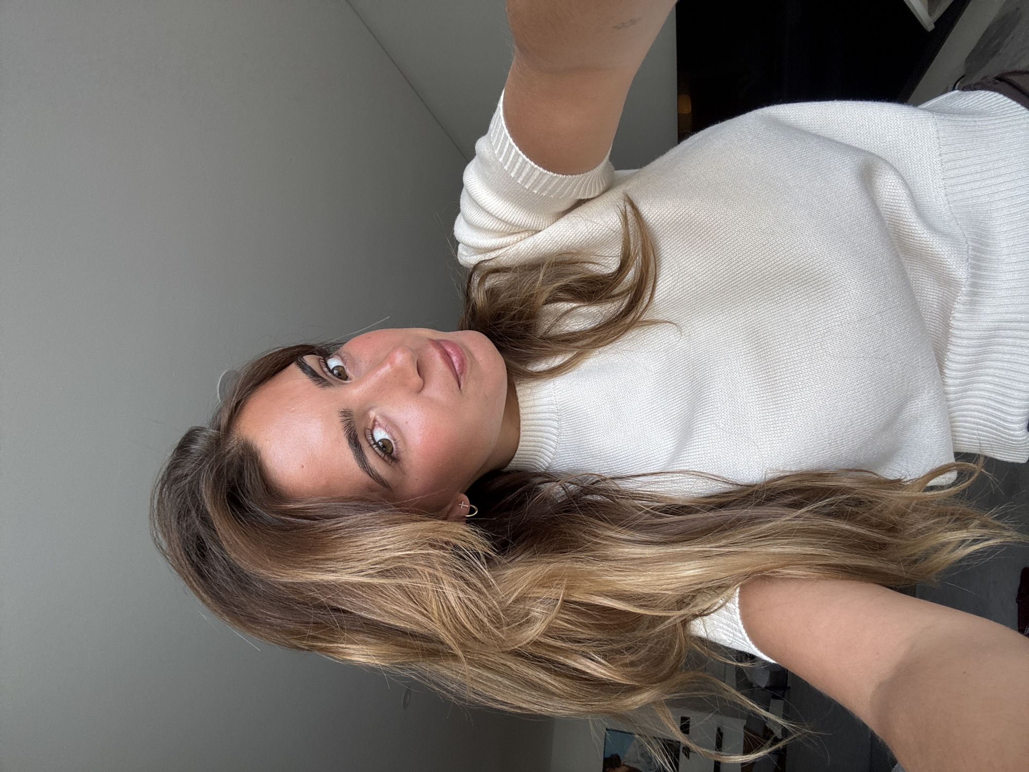 A woman with long, light brown hair is taking a selfie. She is wearing a white sweater. The background includes a grey wall and a glimpse of a dark object. The image appears to be taken indoors. There is no information to suggest the location is Vila Nova de Gaia, Portugal.