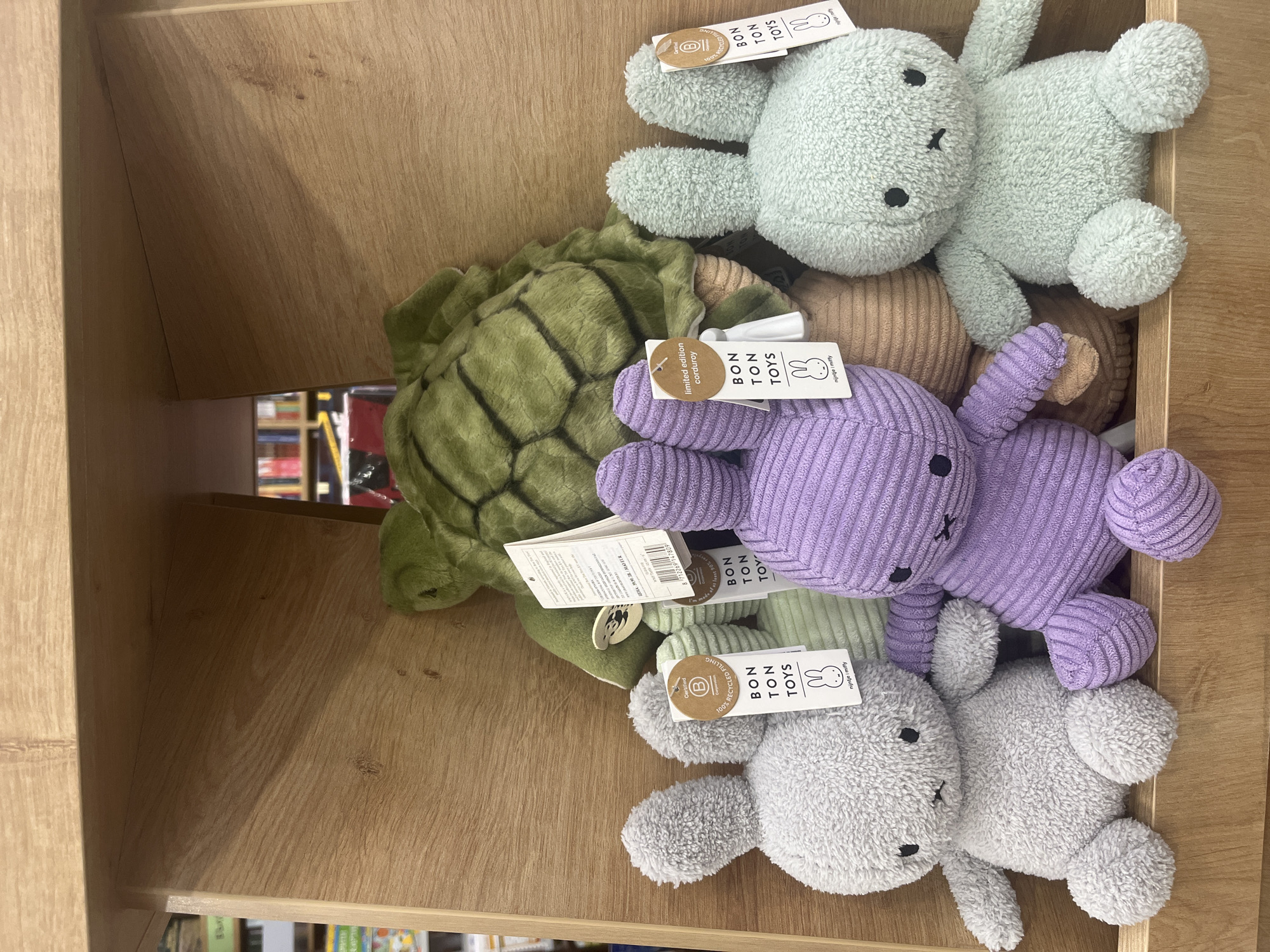 The image shows a shelf in what appears to be a store, filled with various plush toys. The toys are predominantly light green, purple, gray and brown rabbits, and a green turtle. Each toy has a tag attached that displays "BON TON TOYS" and an image of Miffy. The scene does not depict Varna, Bulgaria, nor does it contain information about people, subjects, or actions. The primary focus is solely on the merchandise within the wooden shelf.