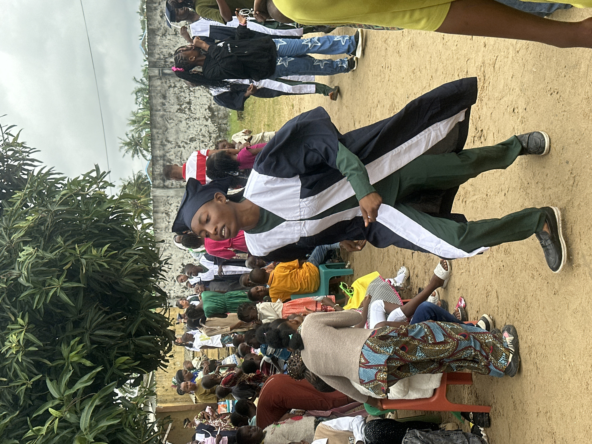 This image captures a gathering of people, likely a graduation ceremony or a school event, set outdoors in Badagry, Nigeria. The scene is dominated by a group of individuals, some wearing academic regalia, including caps and gowns. One person in the foreground is wearing a dark blue and white academic gown over a green garment and dark green pants. They are looking down and appear to be in motion.
Behind them, a crowd of people are seated, some on plastic chairs, others on the ground. Many are dressed in casual attire, with vibrant fabrics and patterns visible, suggesting a festive atmosphere. Children and adults are present, with some children in school uniforms. A large tree with green foliage is on the left side of the frame, providing some shade.
The ground is sandy, and a concrete wall with some greenery and palm trees in the background suggests the location is somewhat rural or a semi-urban area. The sky is overcast, indicating it could be a cloudy day or late afternoon. The overall mood appears to be celebratory and communal. No visible text is present in the image.