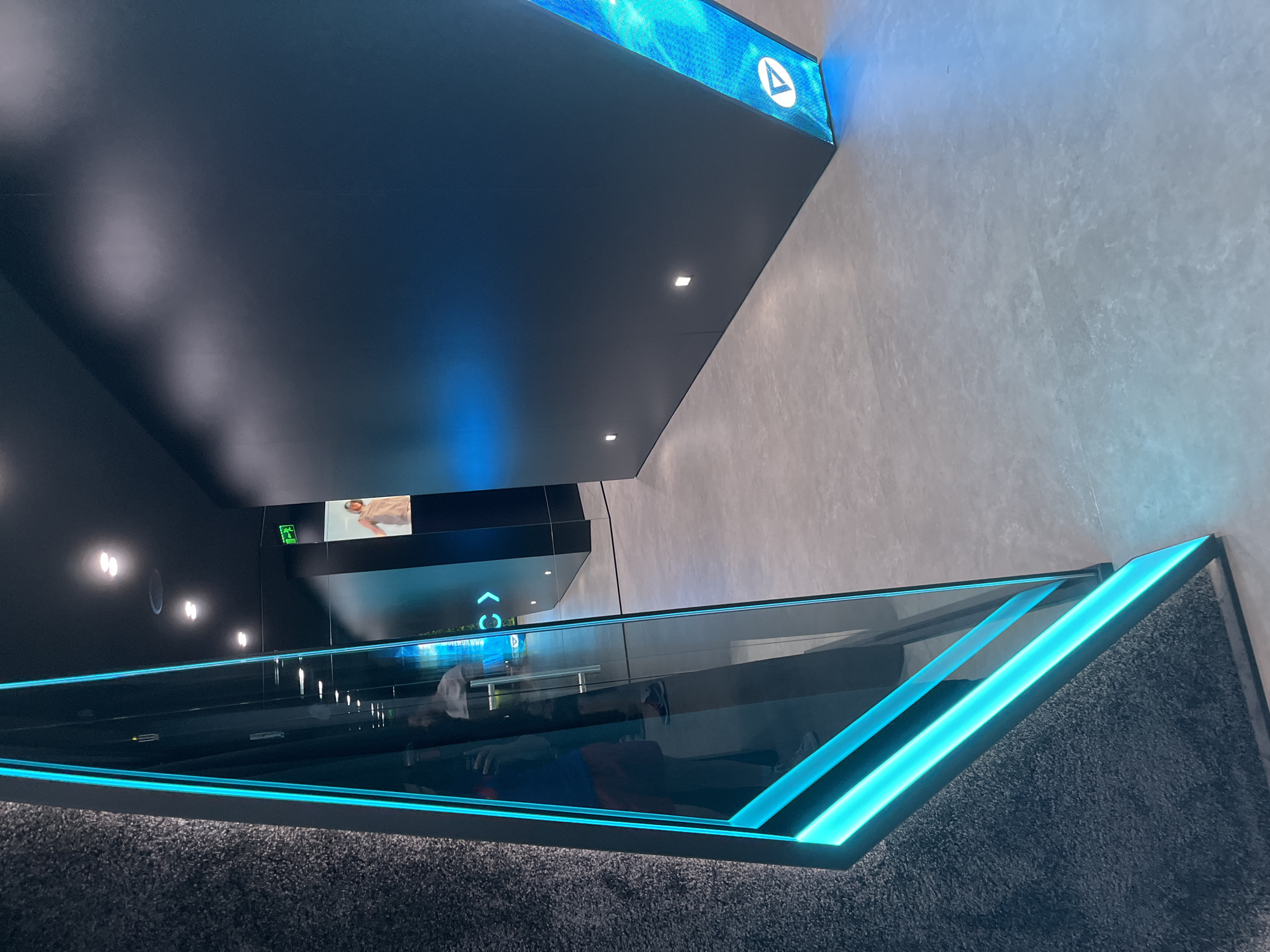 The image captures an interior space with a contemporary design aesthetic. Dominating the upper portion is a large, dark, flat surface extending from the top left corner, punctuated by recessed lighting. The ceiling structure features a blue-lit panel displaying a logo, contrasting with the muted tones of the wall to its right.
Below this, a transparent barrier with vibrant blue LED lighting runs diagonally across the frame. Reflections visible within the barrier suggest its glass construction. The area beneath is covered by a dark, textured carpet. The space is well-lit, with additional light sources visible further in the distance. Overall, the scene conveys a sense of modernity and sophistication. The city of Unknown and the specific location within Unknown are not discernible from the image.