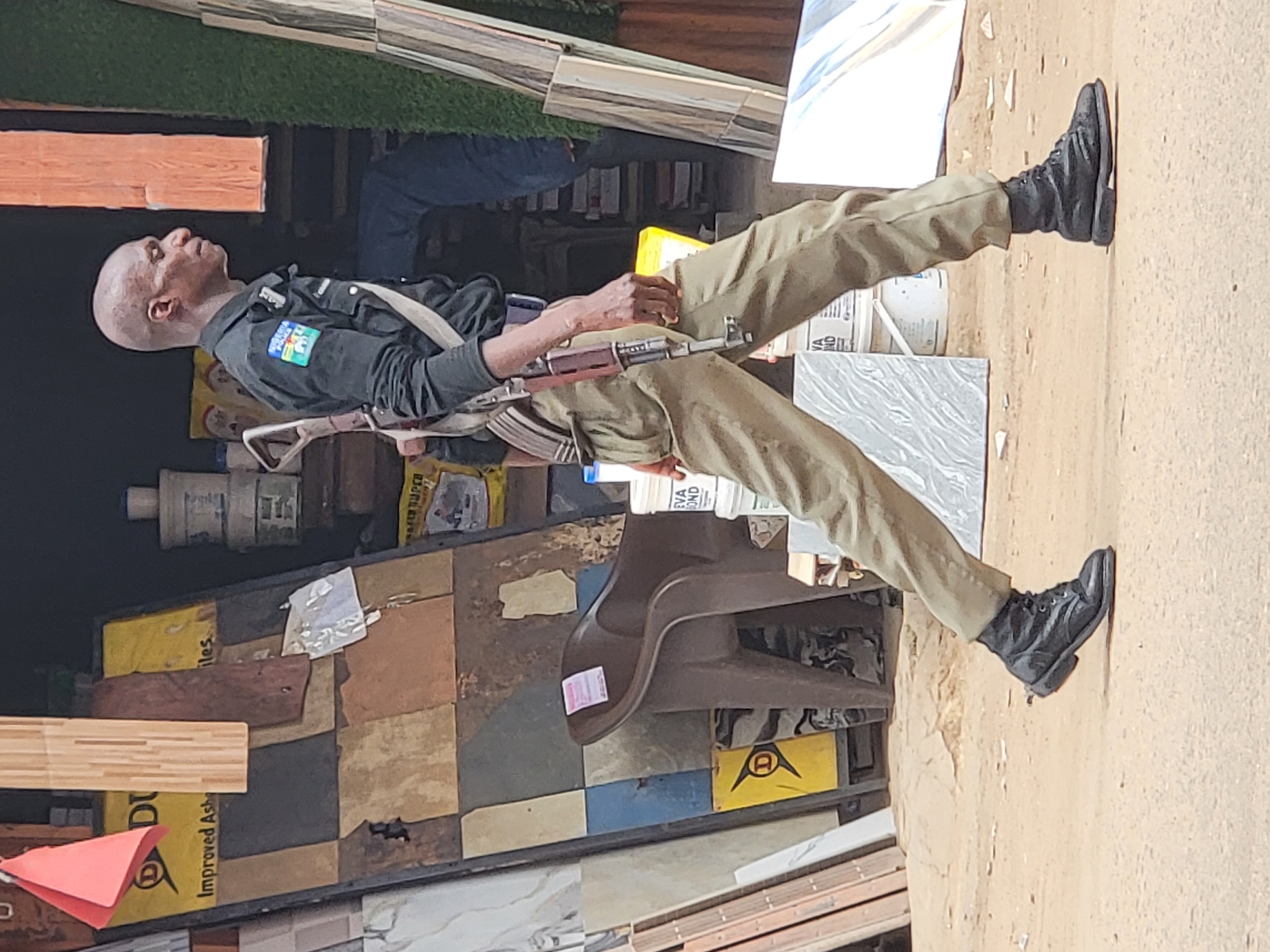 A man, likely a Nigerian police officer, dressed in uniform, complete with an AK-47 slung across his torso, stands on what appears to be a display of tiles and construction materials. The location is suggested as Maiburiji, Nigeria. He is wearing black boots.